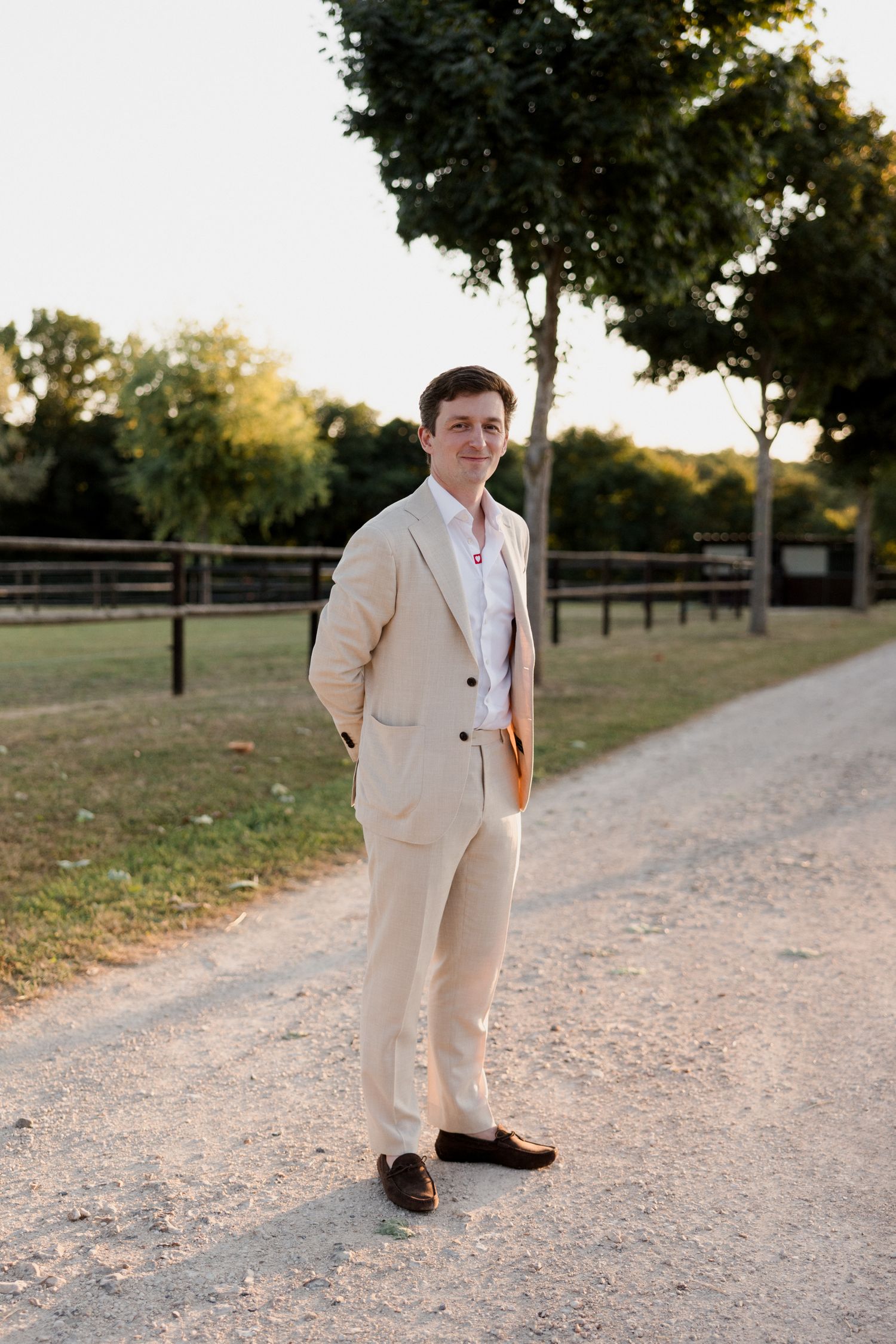 A figure in a light beige suit walks along a gravel path surrounded by trees during golden hour lighting. Photographe mariage Tours