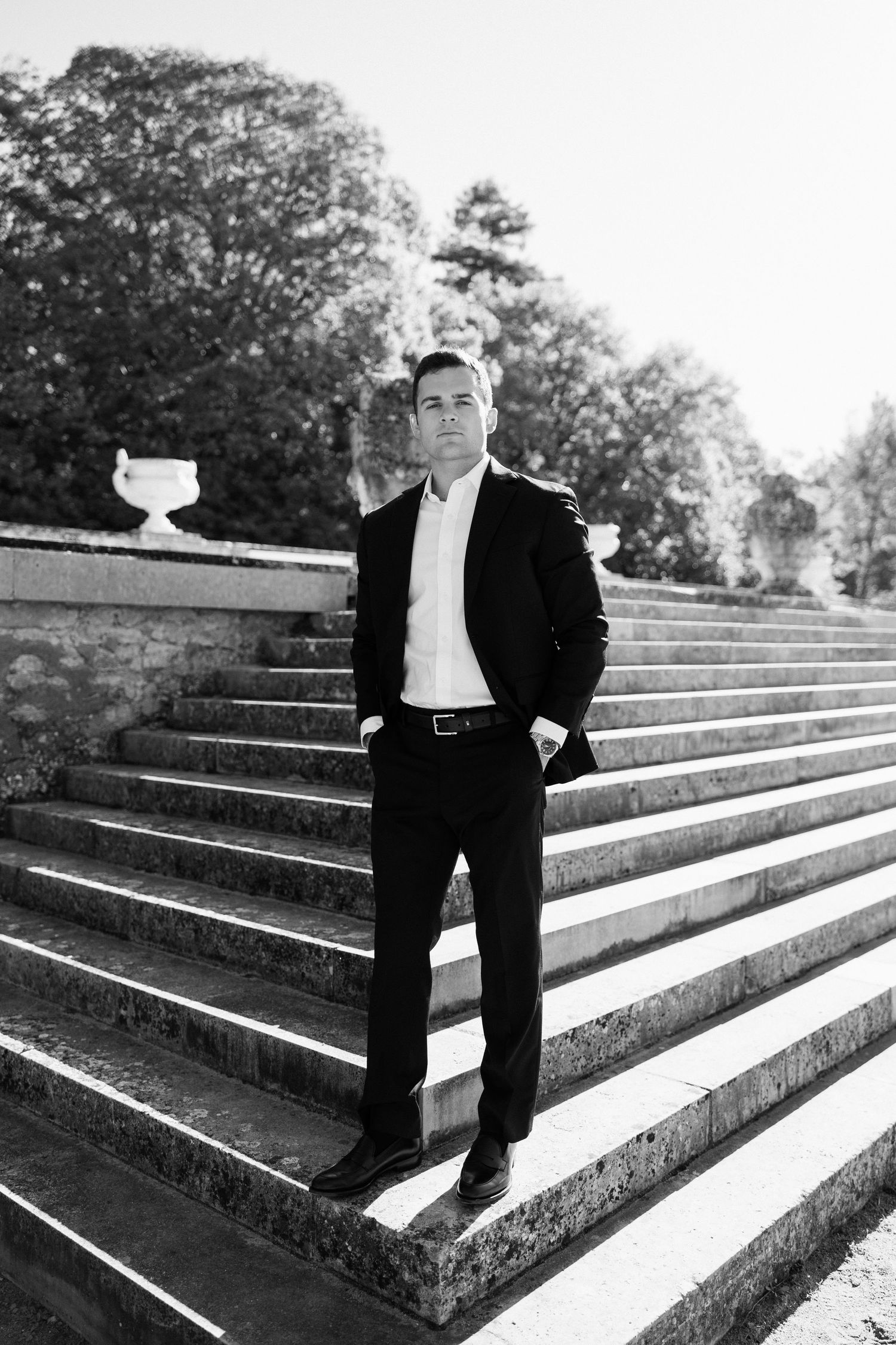 Elegant groom in a black suit stands confidently on stone steps in a classic black and white portrait.