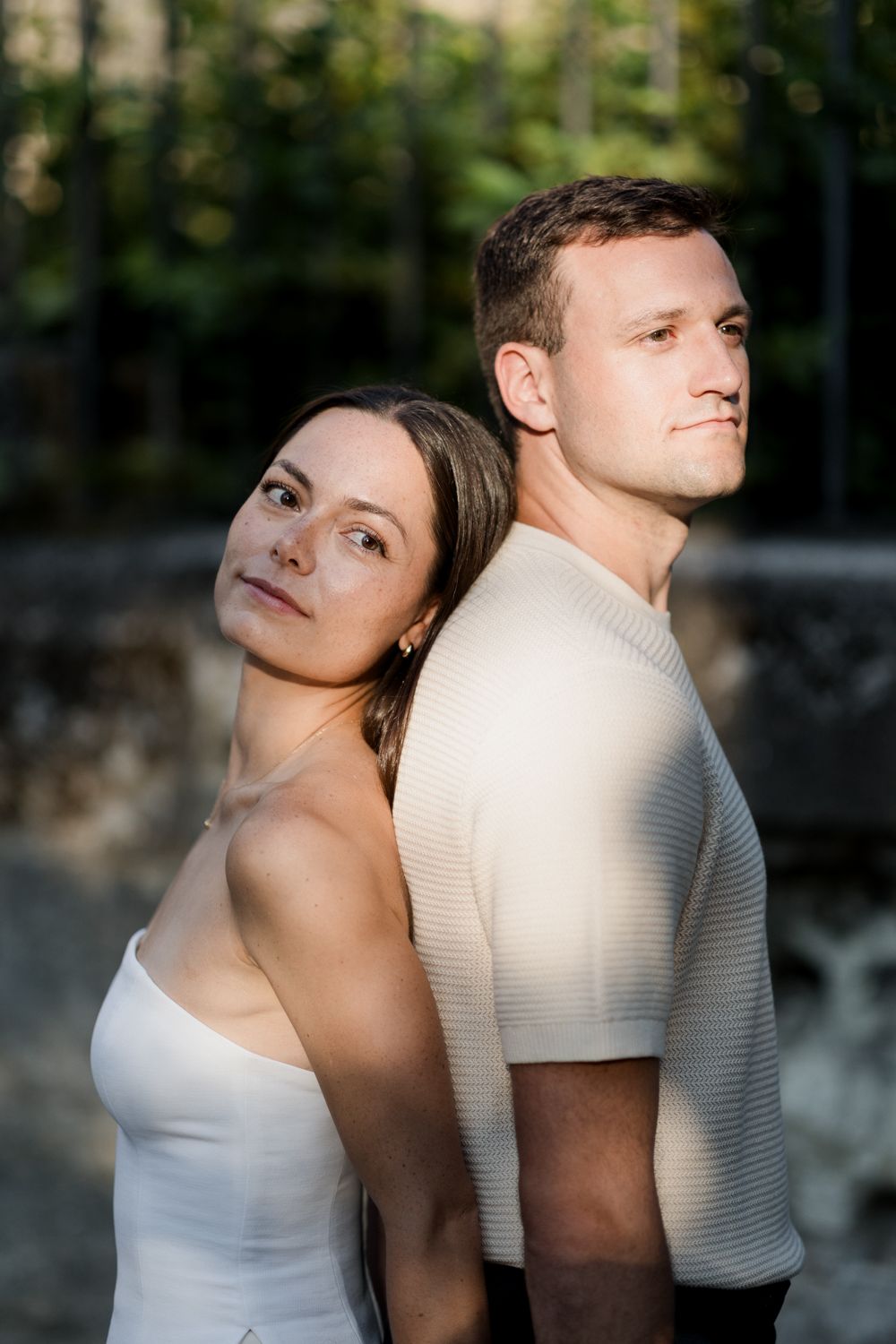 A romantic portrait of a couple standing back to back in white outfits against a stone wall background in natural lighting. Séance couple