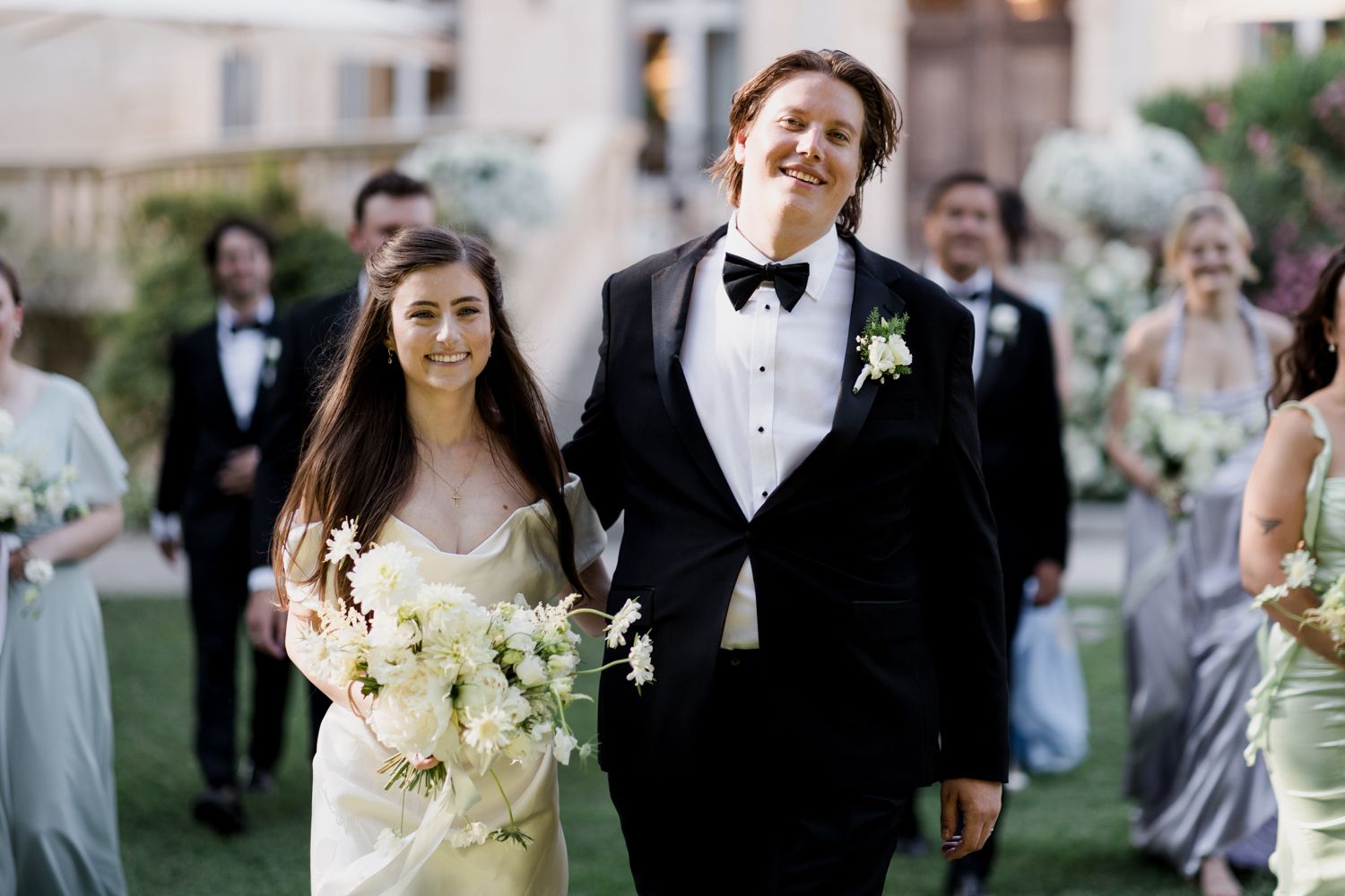 A couple walks together during an outdoor wedding ceremony, the bride holding light-colored flowers and the groom in a tuxedo.