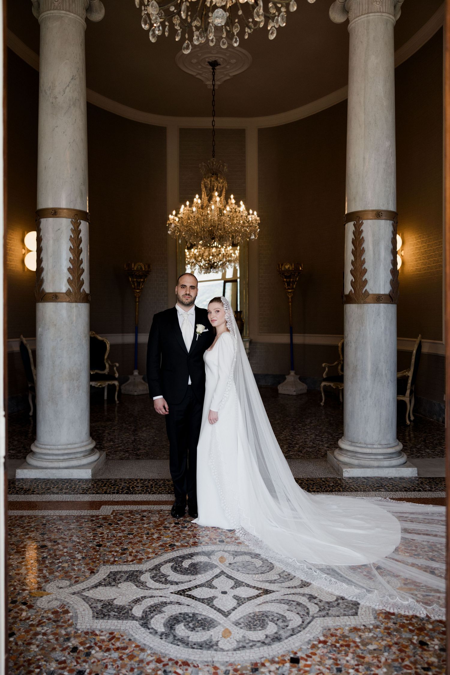 A bride and groom stand in an elegant ballroom with ornate columns and a decorative mosaic floor pattern beneath chandeliers.