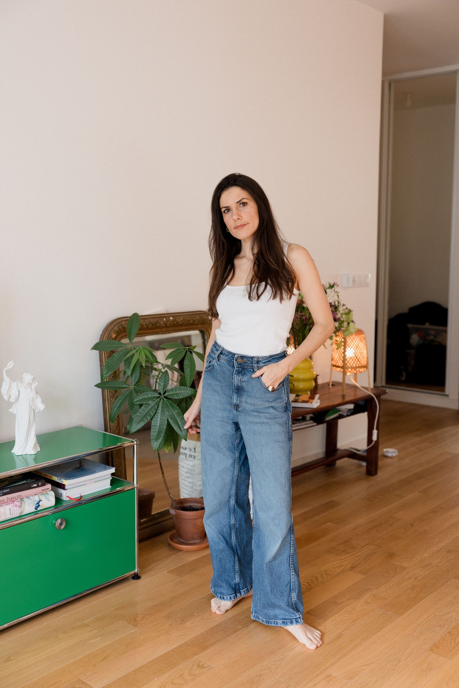 Woman in white tank top and wide-leg blue jeans standing barefoot in a modern living room with green furniture.