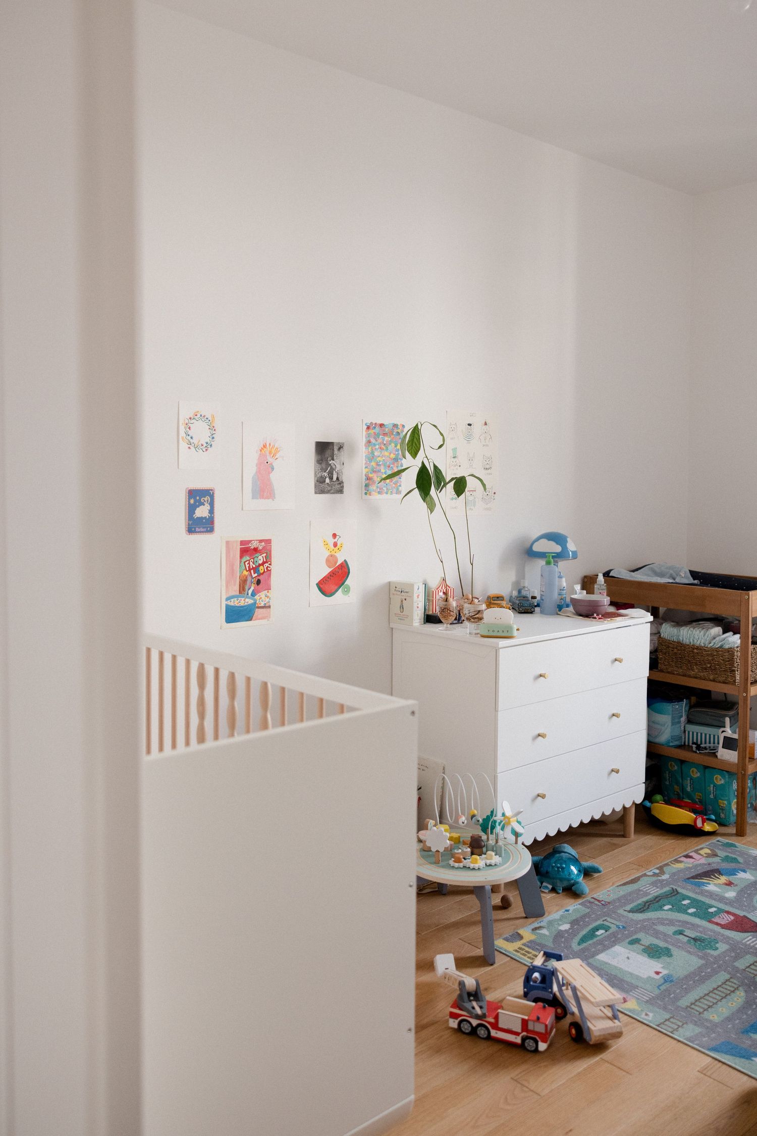 A bright white nursery with a wooden crib, dresser, colorful wall art, potted plant, and children's toys on hardwood floors.