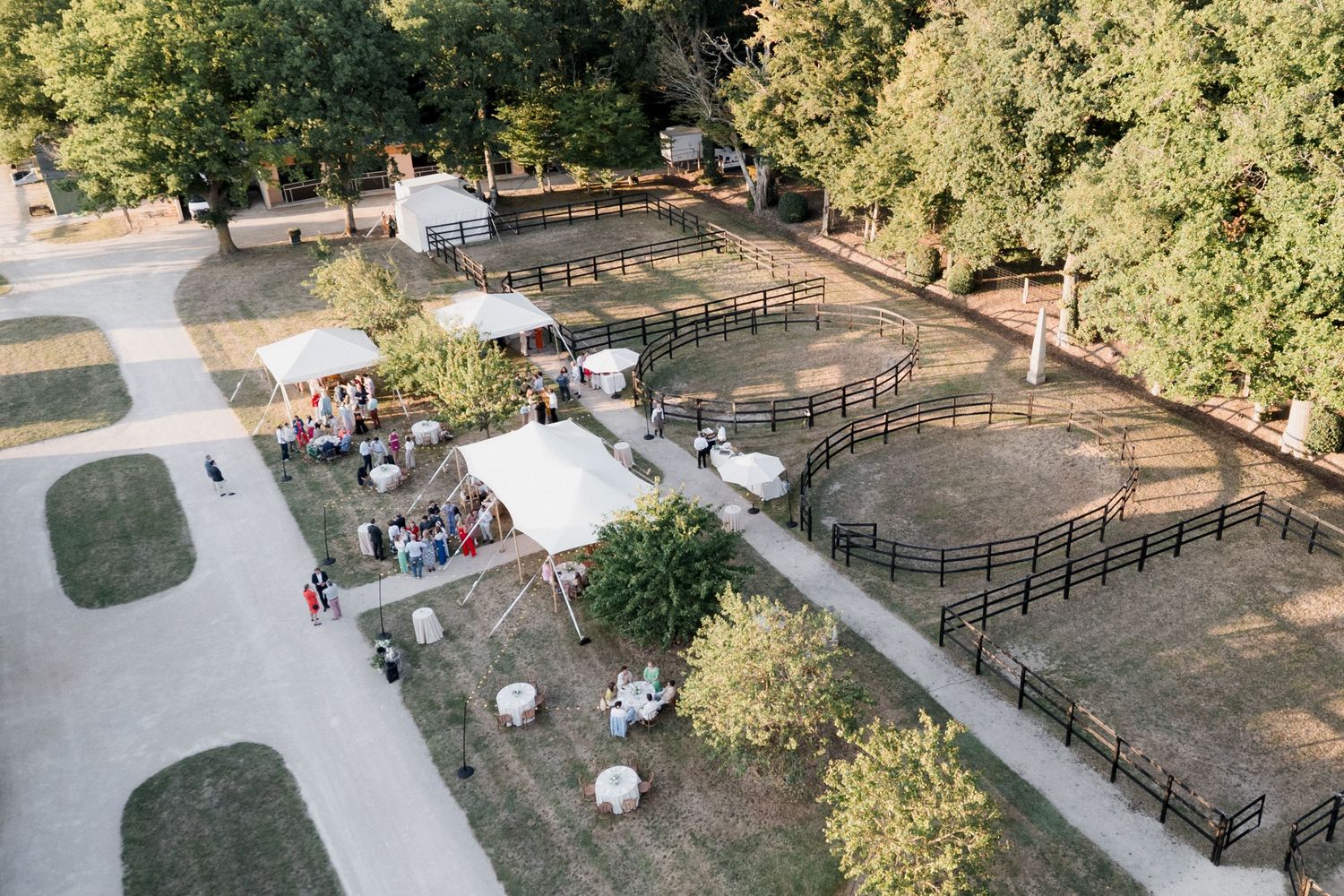 Aerial view of a horse arena surrounded by wooden fencing and trees with riders practicing during daytime.