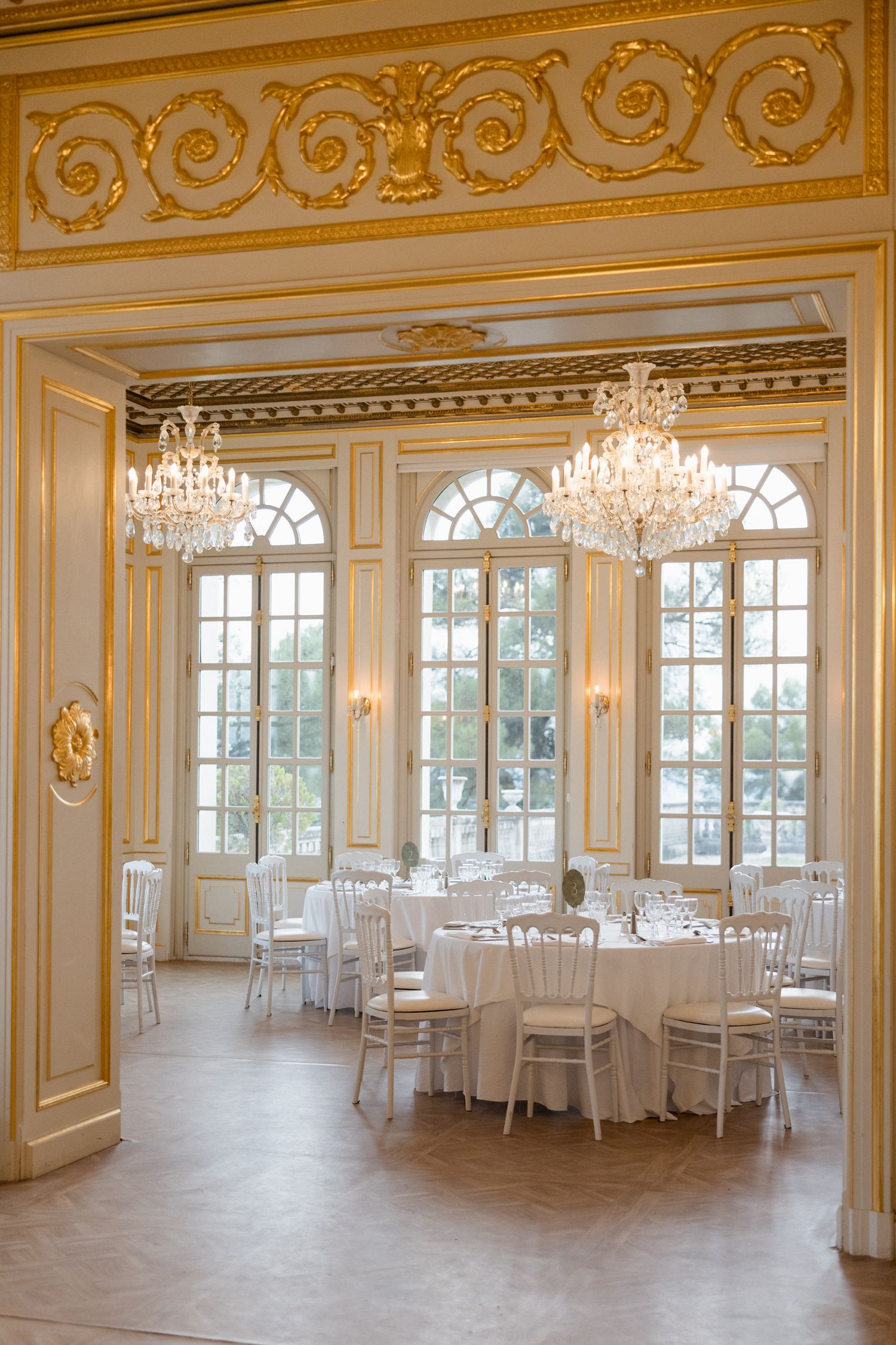 Elegant ballroom with ornate gold trim, crystal chandeliers, arched windows and white round tables set for a formal event. mariage grasse