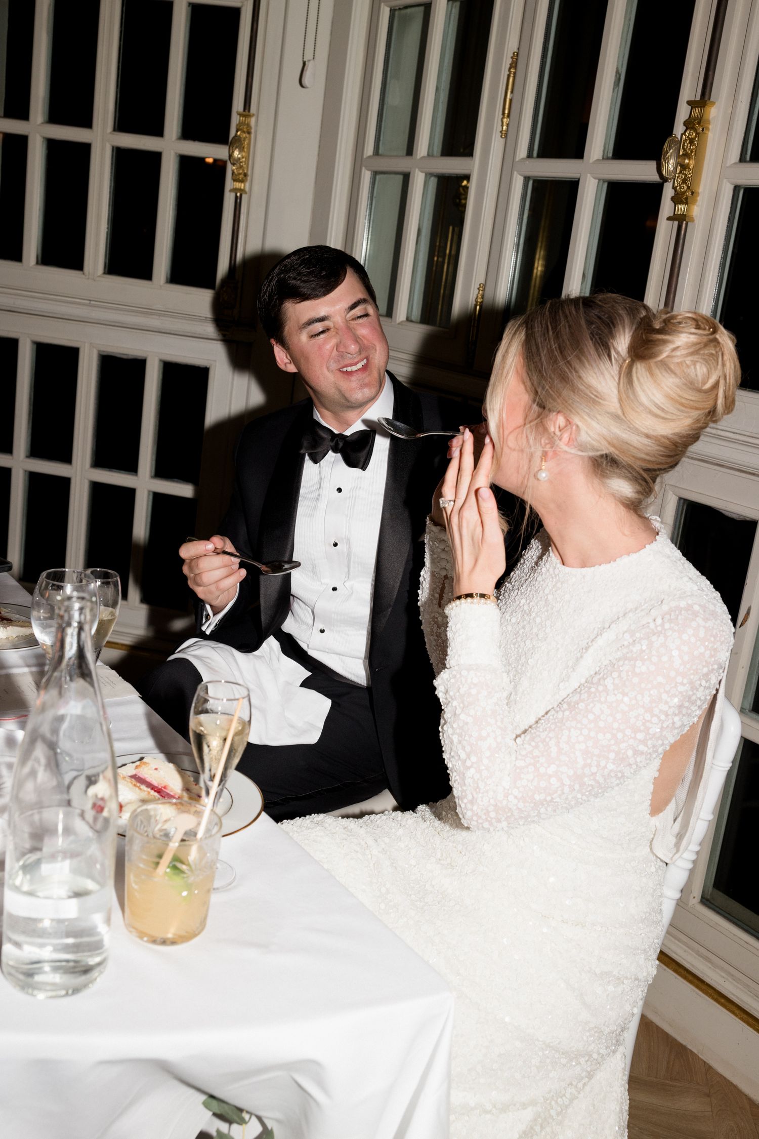Couple in formal attire share laughter at an elegant dinner table with champagne glasses and white tablecloth. Photographe mariage france