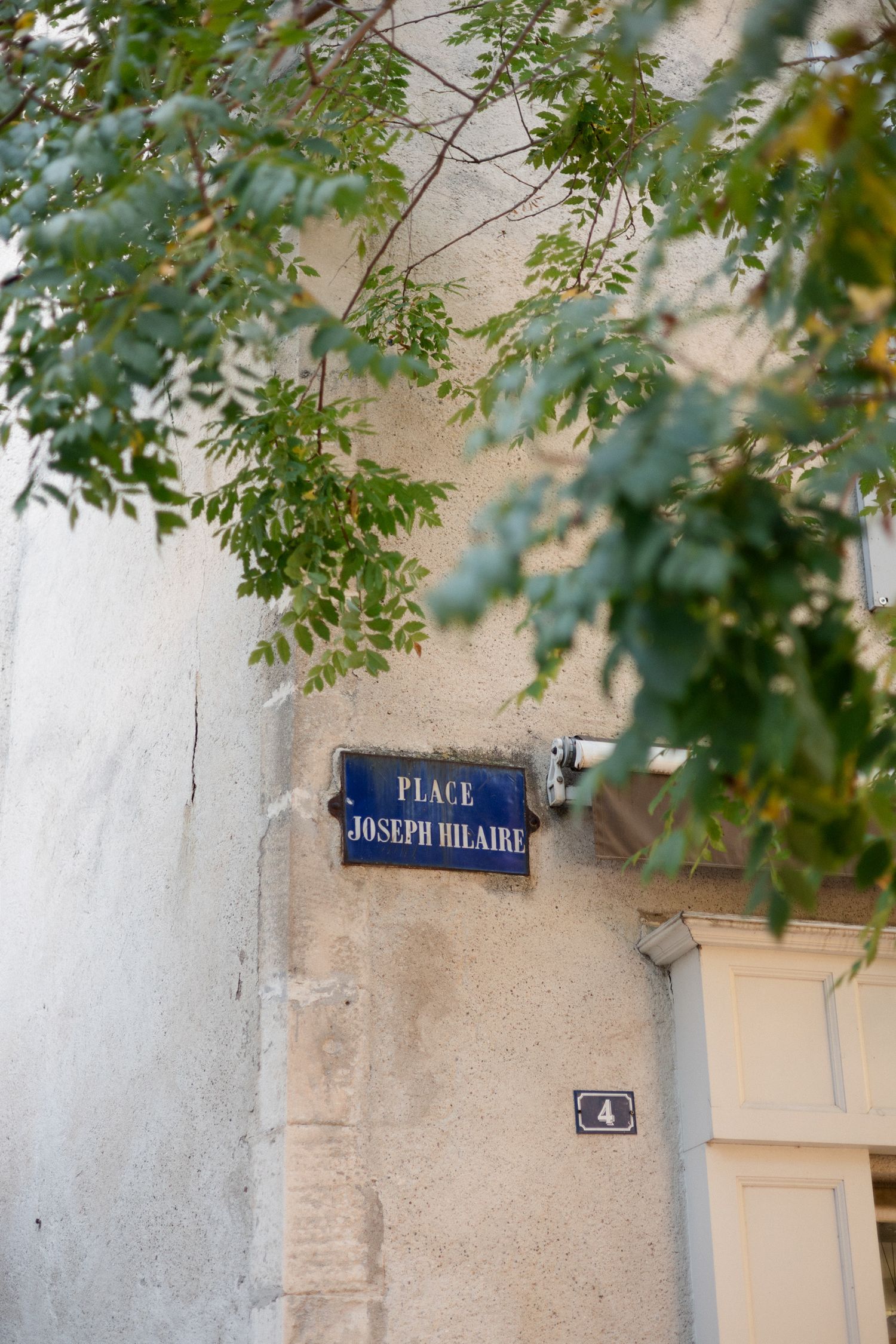 A French street sign reading 'Place Joseph Hilaire' mounted on a light-colored wall with green leaves overhead.