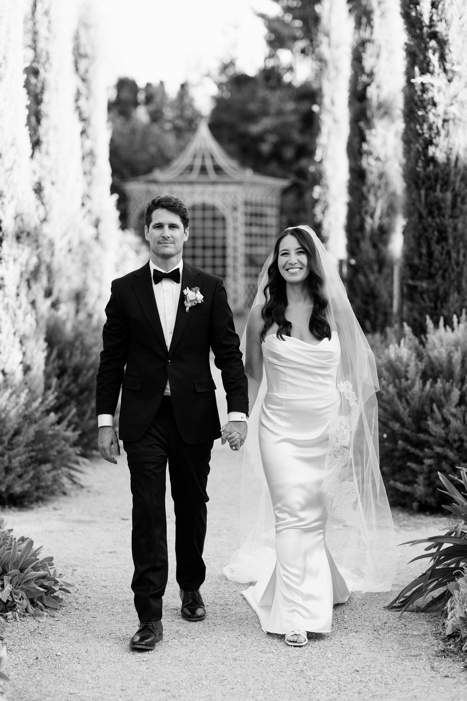 Black and white triptych of wedding couple walking through a cypress tree-lined path at an outdoor venue.