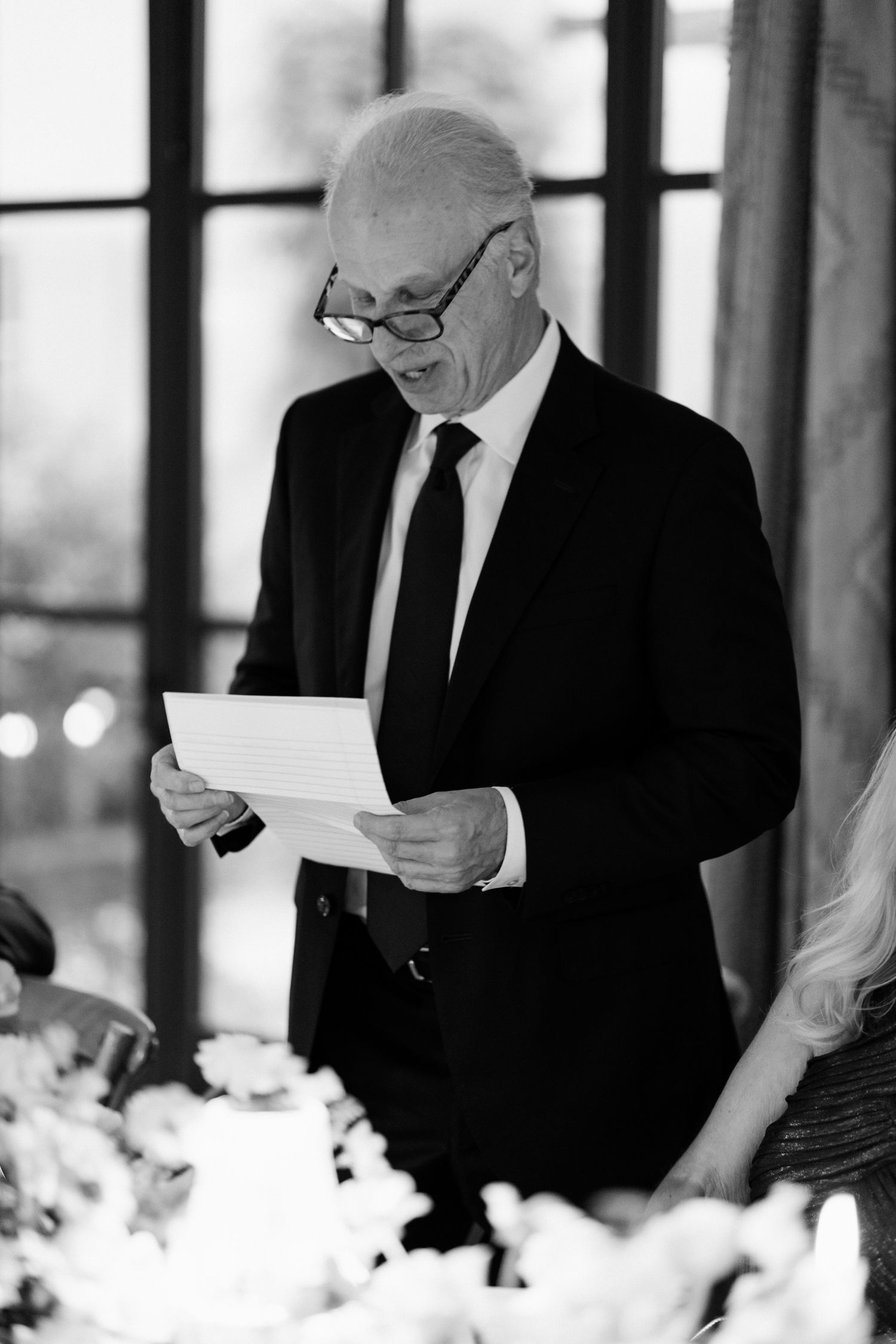 Black and white photo of people reading papers during a formal event with white floral decorations.