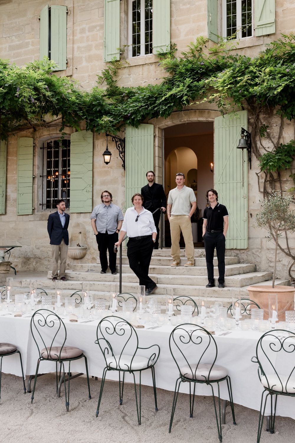 A French country house exterior with mint green shutters, climbing vines, and decorative iron chairs on the patio. Auberge de la Treille