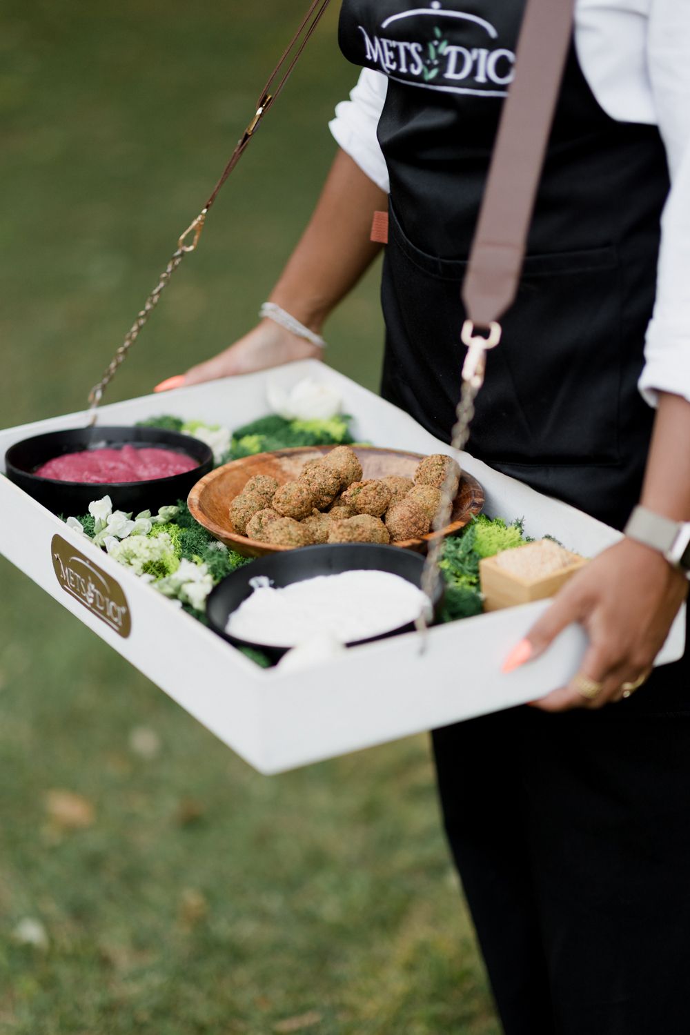 Server in black apron holds white takeout container with fresh salad and bread against grassy outdoor background. Mets d'ici
