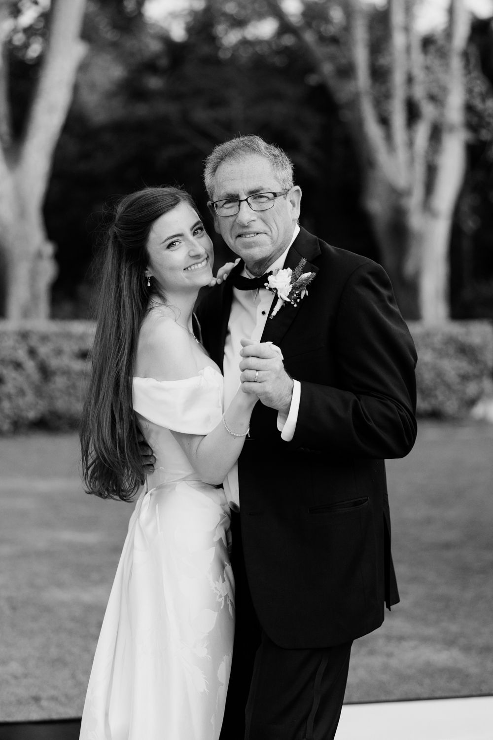 A bride in white dress dances with a man in black tuxedo during a tender outdoor wedding moment.