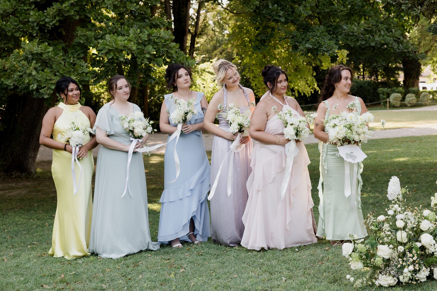 A group of bridesmaids wearing pastel dresses with flowers stands together outdoors at a garden wedding.