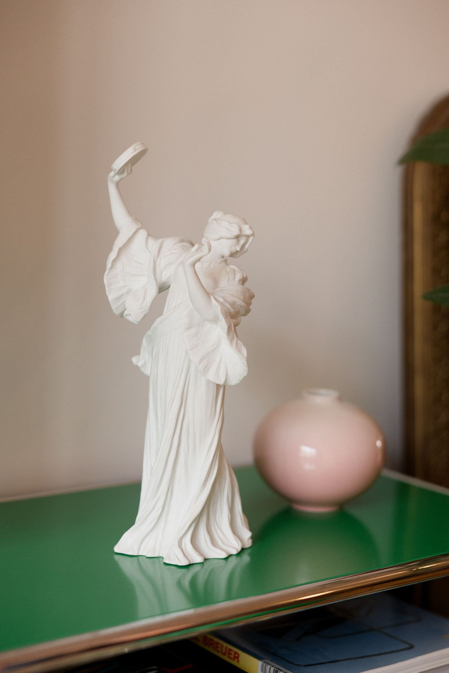 White porcelain female figurine beside a pink ceramic vase on a green glass shelf.