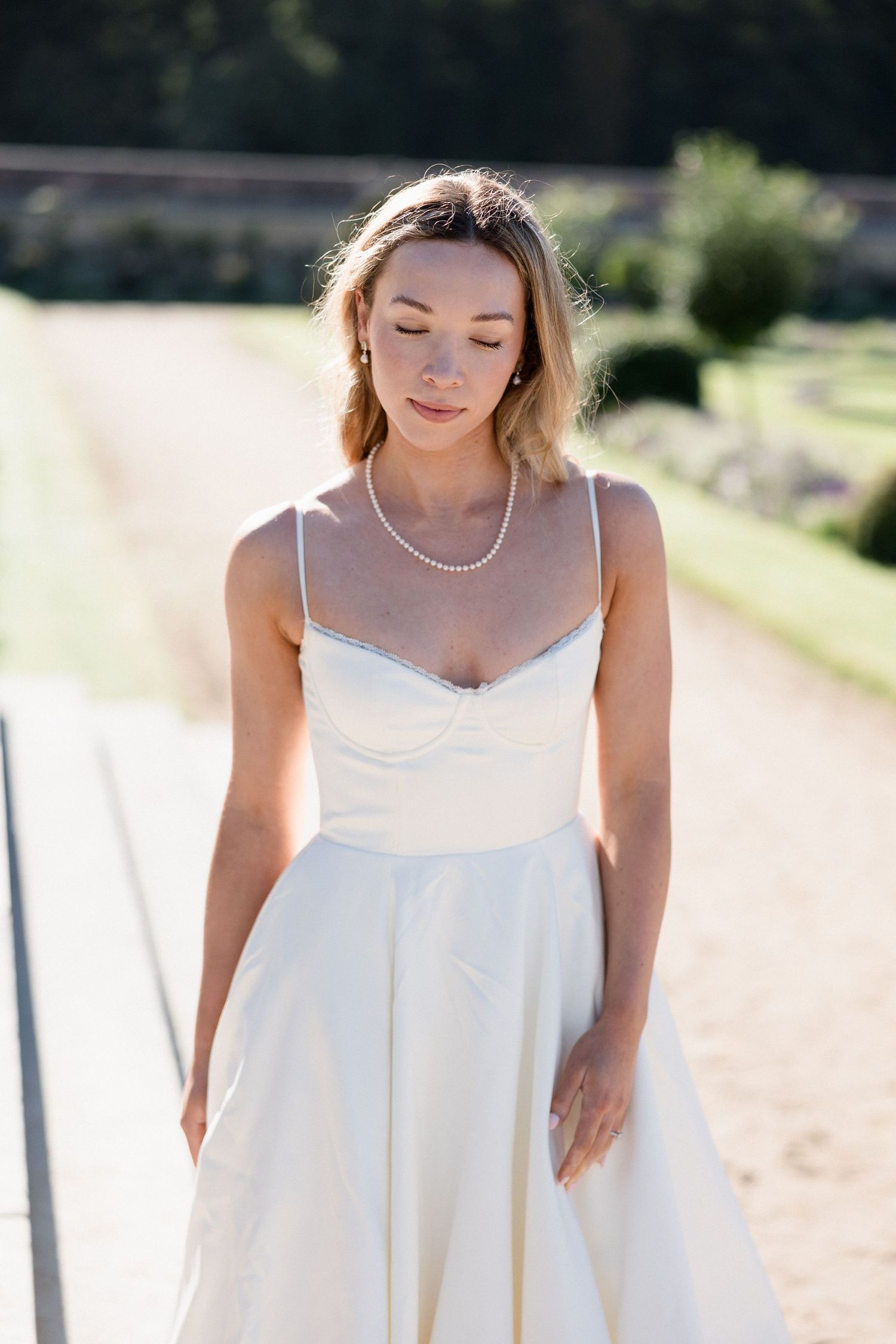 Blonde bride in satin slip gown with pearl necklace poses outdoors in warm sunlight.