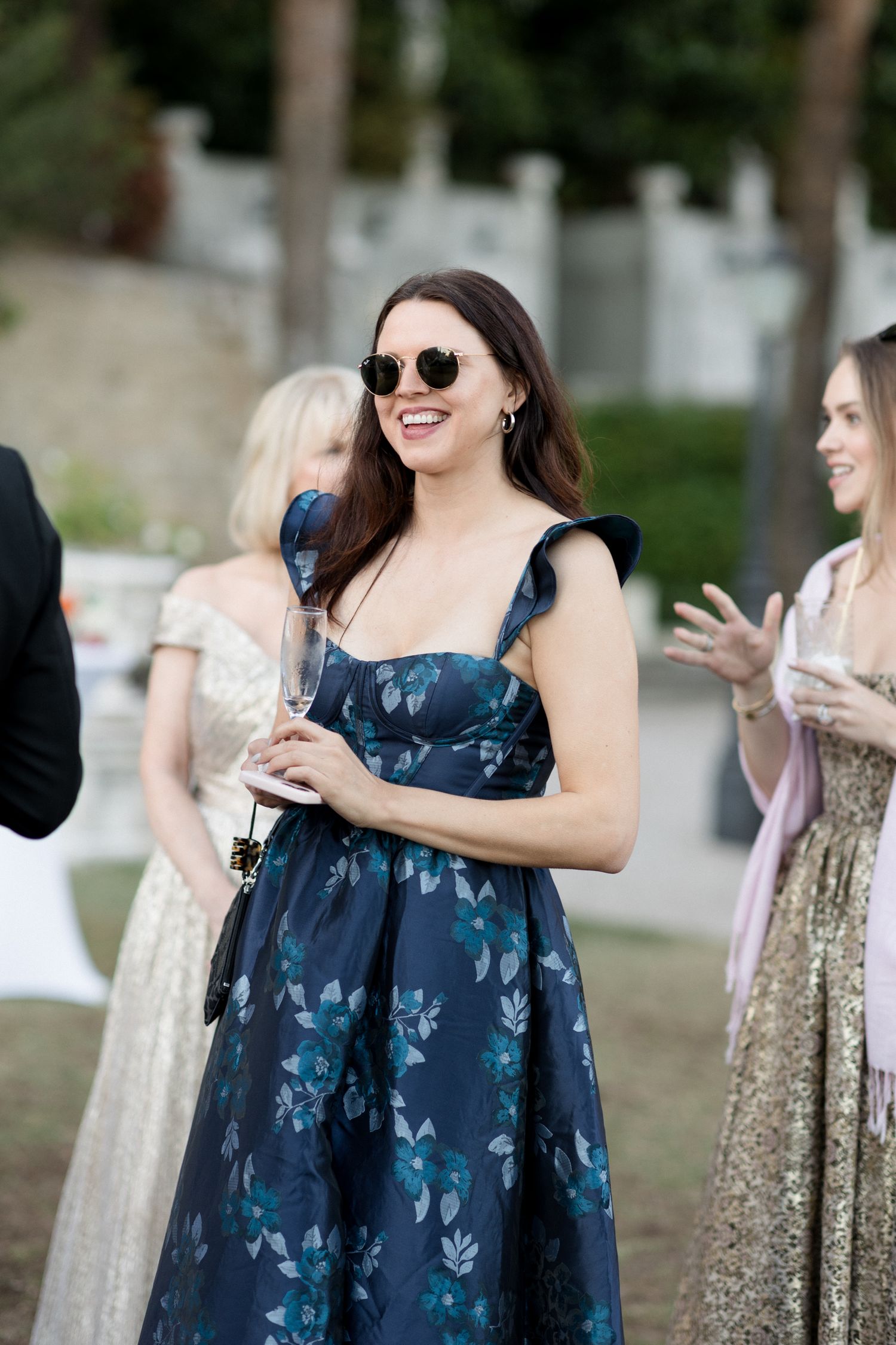 Woman in navy floral dress and sunglasses smiles while holding a drink at an outdoor event.