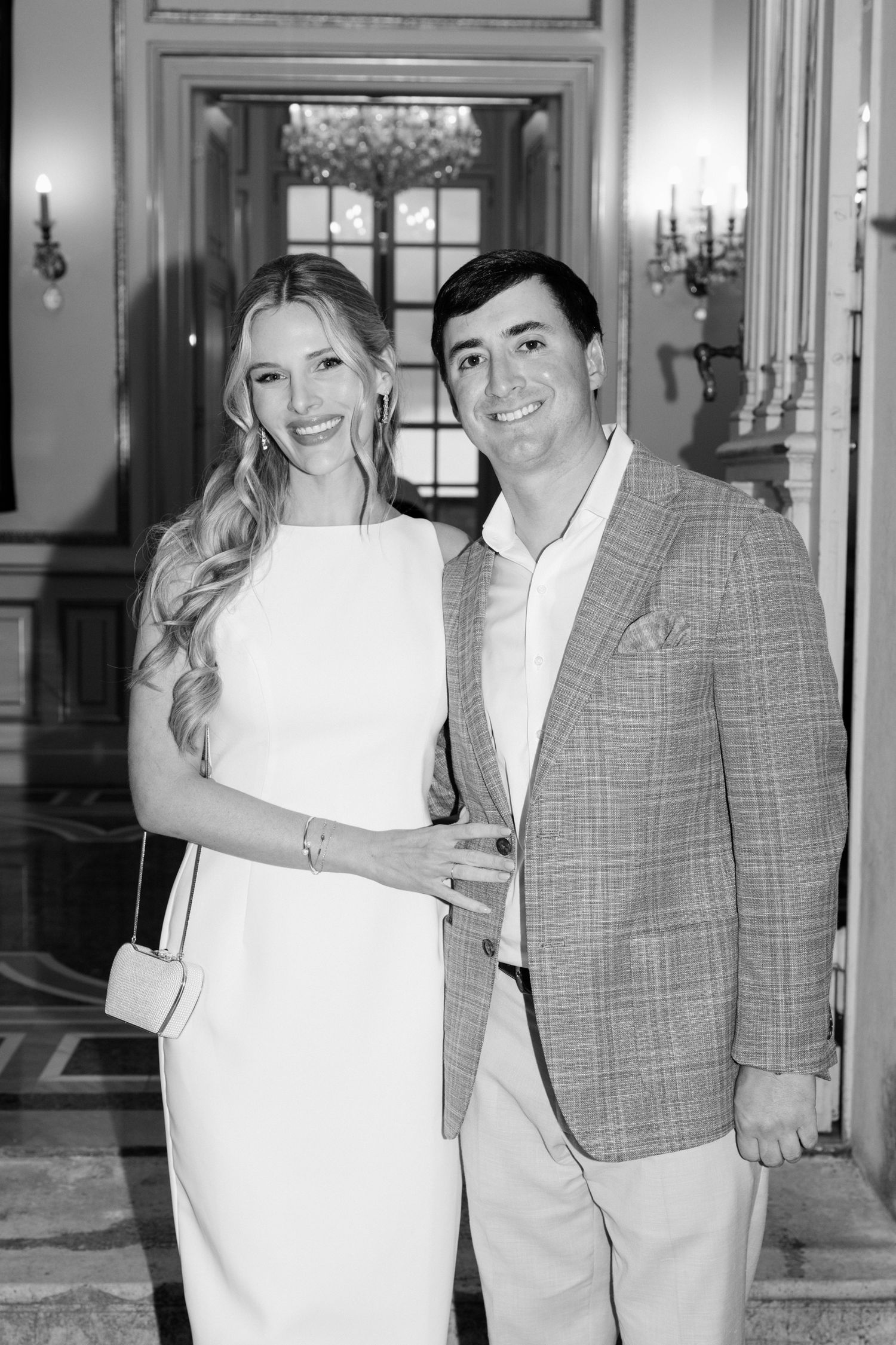A couple poses together in formal attire, with a woman in white and a man wearing a plaid blazer.