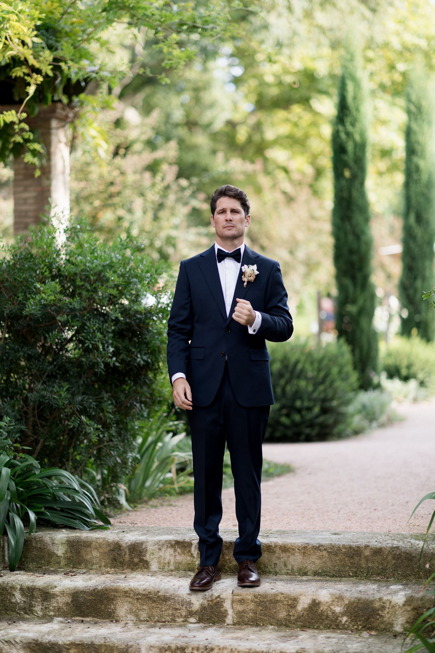A person in a classic black tuxedo and bowtie stands on stone steps in a lush garden setting. Editorial wedding photographer france