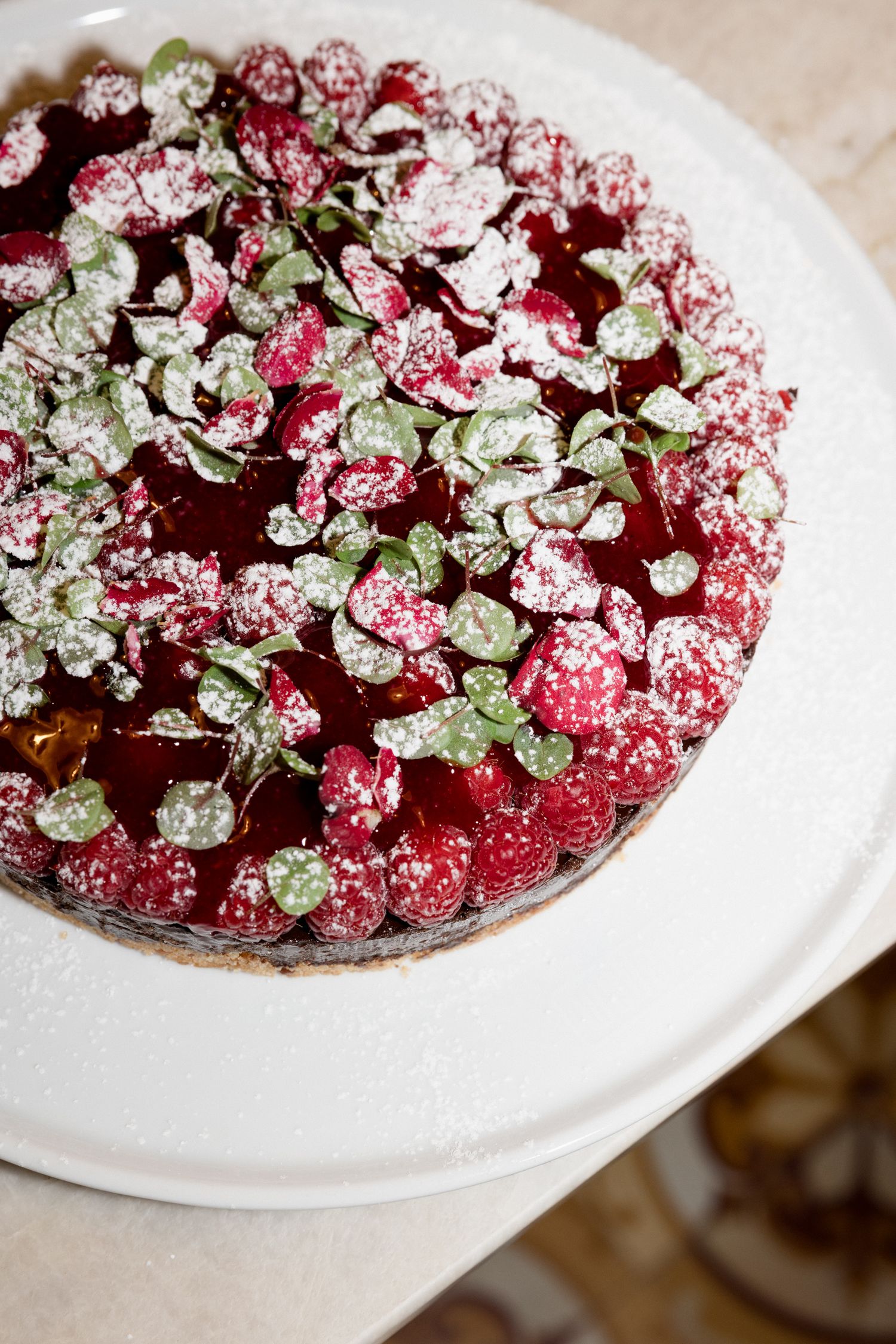 Closeup view of a chocolate raspberry tart garnished with fresh mint leaves and powdered sugar on a white plate.
