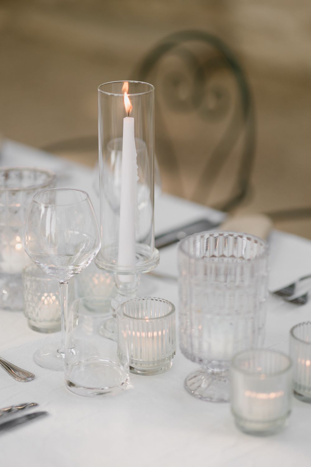 Elegant table setting with white candle in glass hurricane, crystal glasses and votives on white tablecloth.