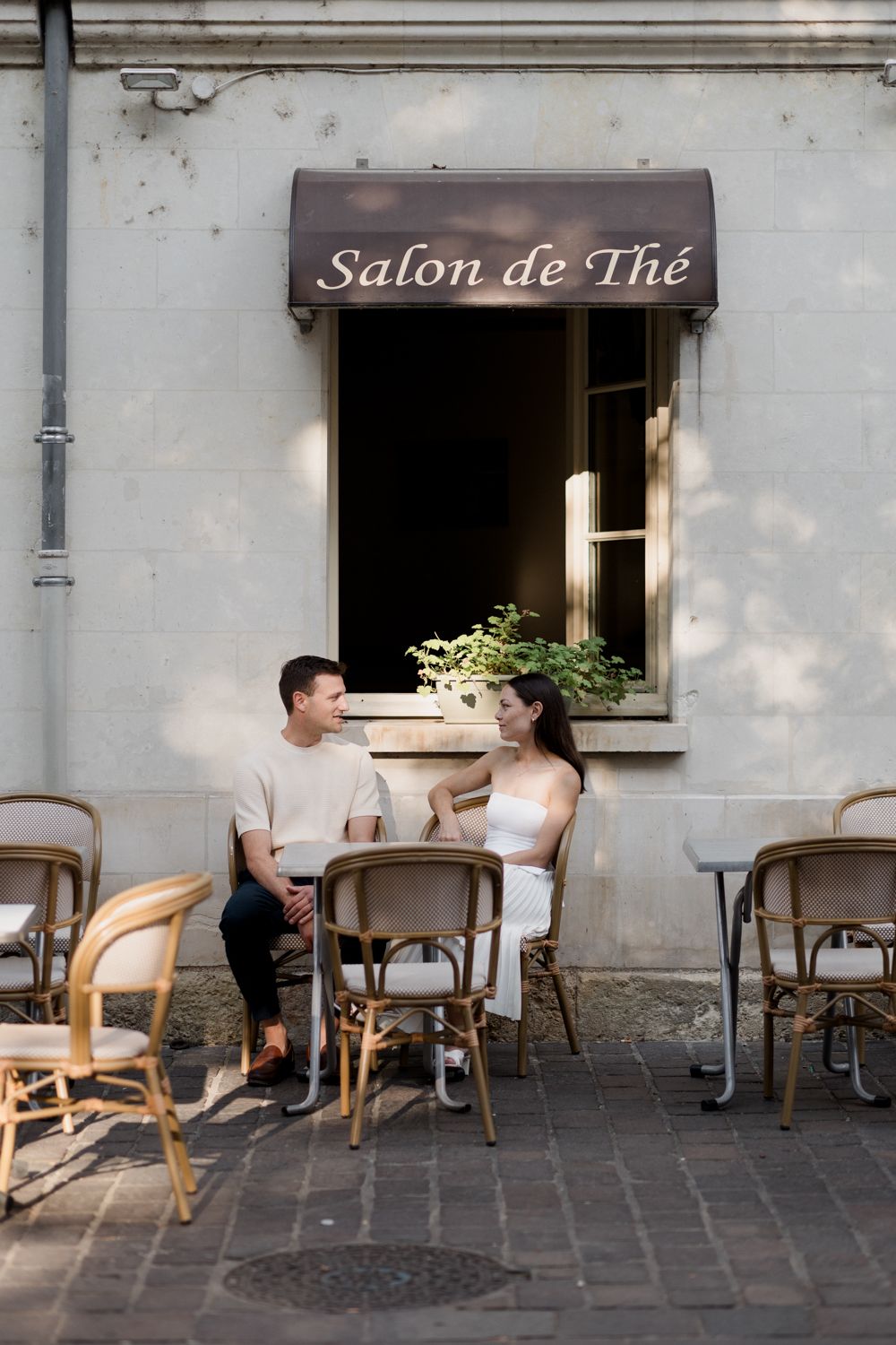 A romantic couple enjoys conversation at an outdoor Salon de The cafe with bentwood chairs and stone paving.