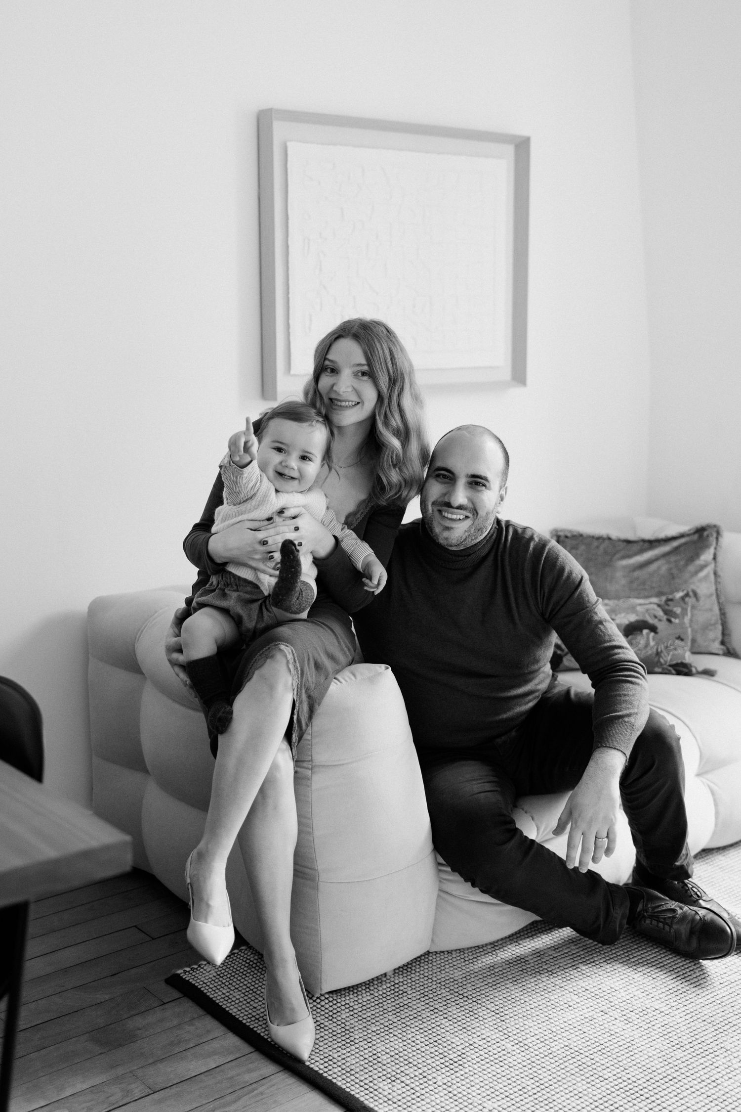 Black and white family portrait of smiling parents with baby sitting on sofa in modern living room.