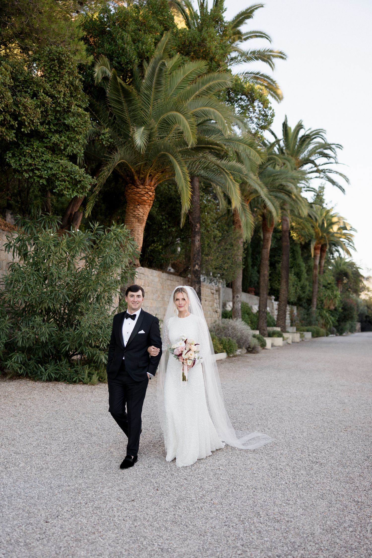 Wedding couple walks together on a scenic palm-lined gravel pathway with stone walls at dusk. Photographe mariage france grasse cote d'azur