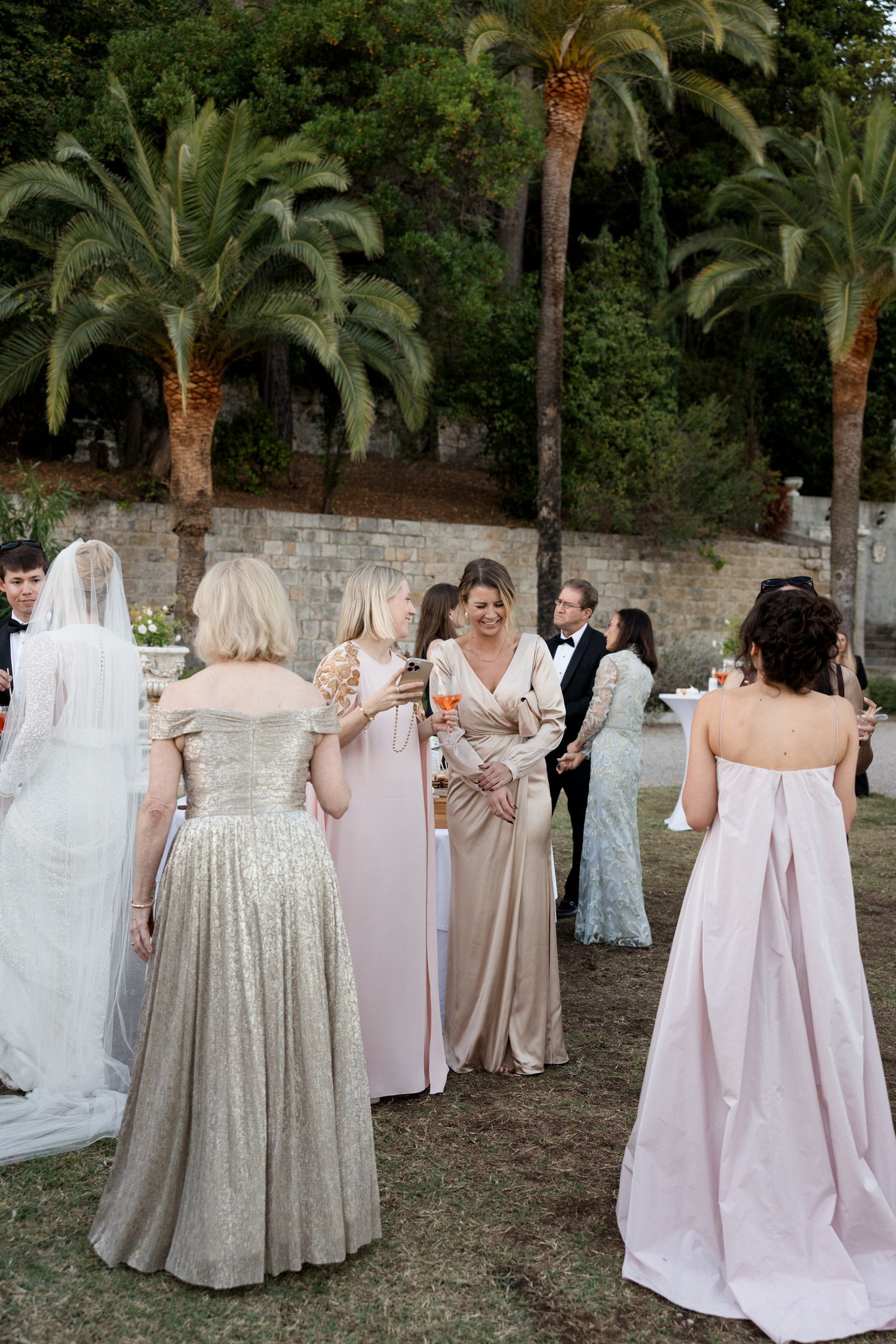 Wedding guests in formal attire gather outdoors among palm trees at an elegant evening celebration.