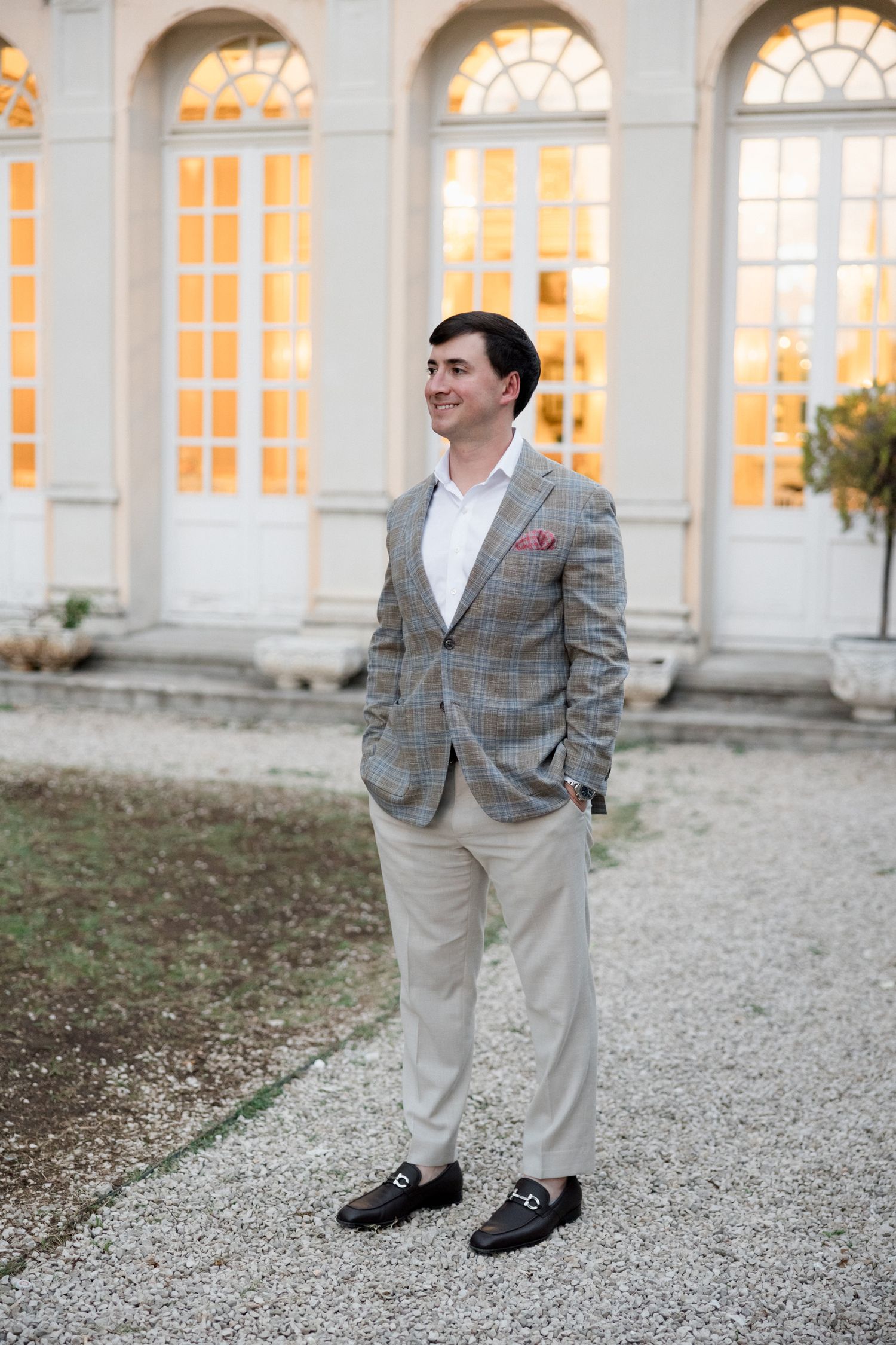Groom in grey plaid blazer and khaki pants poses on gravel path outside ornate building with arched windows.