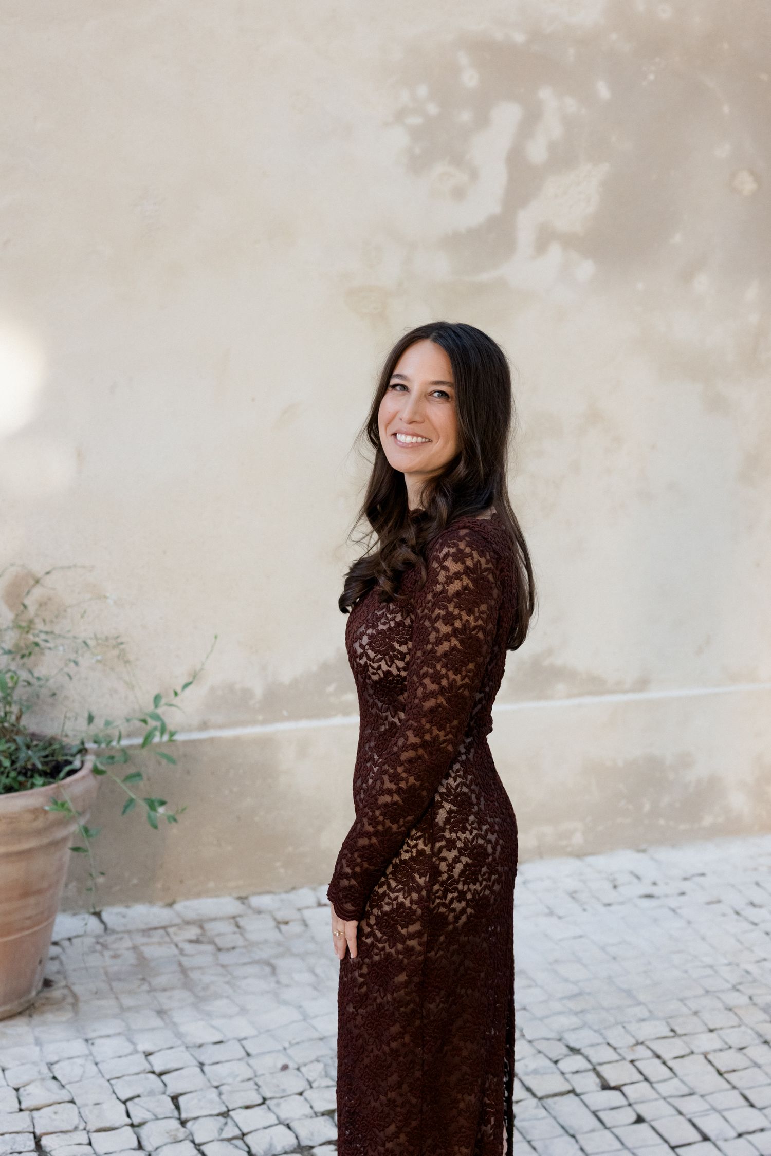 Woman in elegant brown lace dress poses against textured wall with potted plants and cobblestone ground. Engagement session Paris