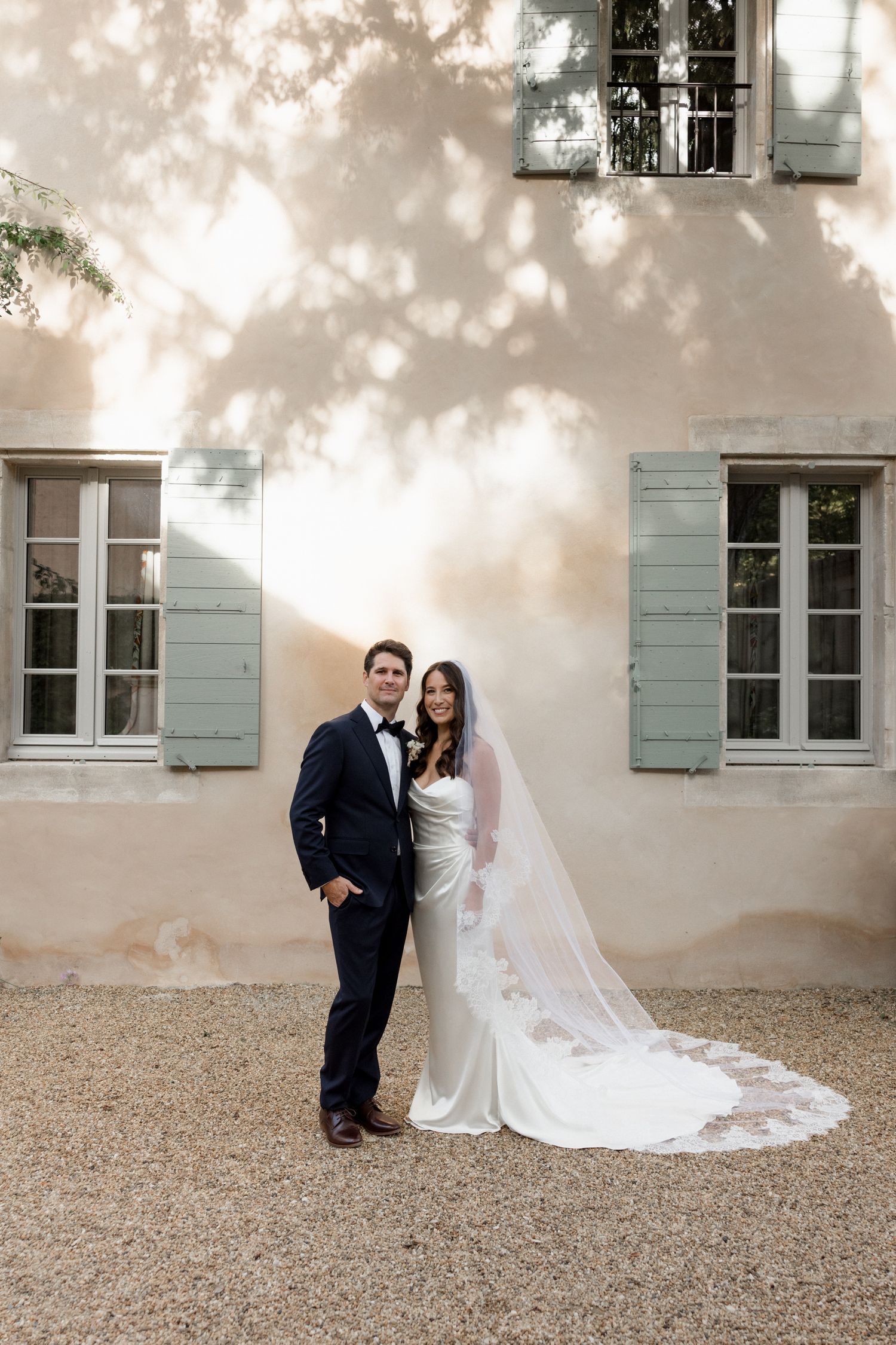 A romantic wedding couple poses against a light colored building with green shuttered windows and gravel ground. wedding photographer france