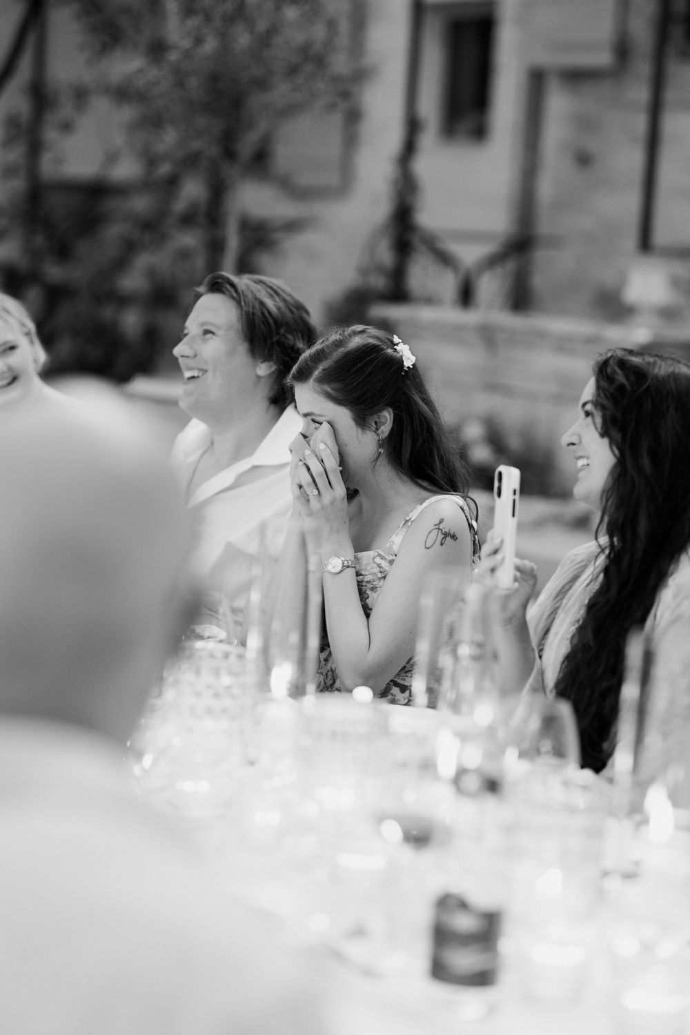 Black and white photo of guests laughing during wedding speeches at reception table with glassware.
