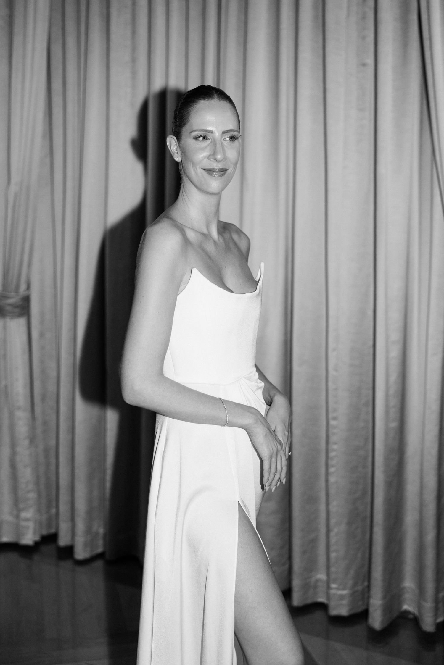 Black and white photo of a woman in a white spaghetti strap gown with a slit, posing in front of draped curtains.