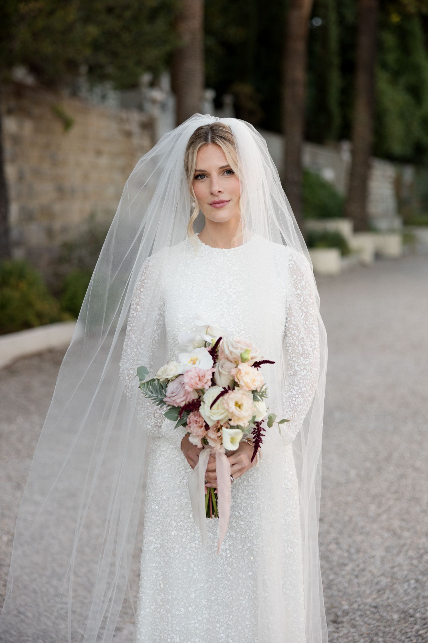 A bride in a long-sleeved white lace wedding dress holds a soft pink and white bouquet on a tree-lined path. Photographe mariage france