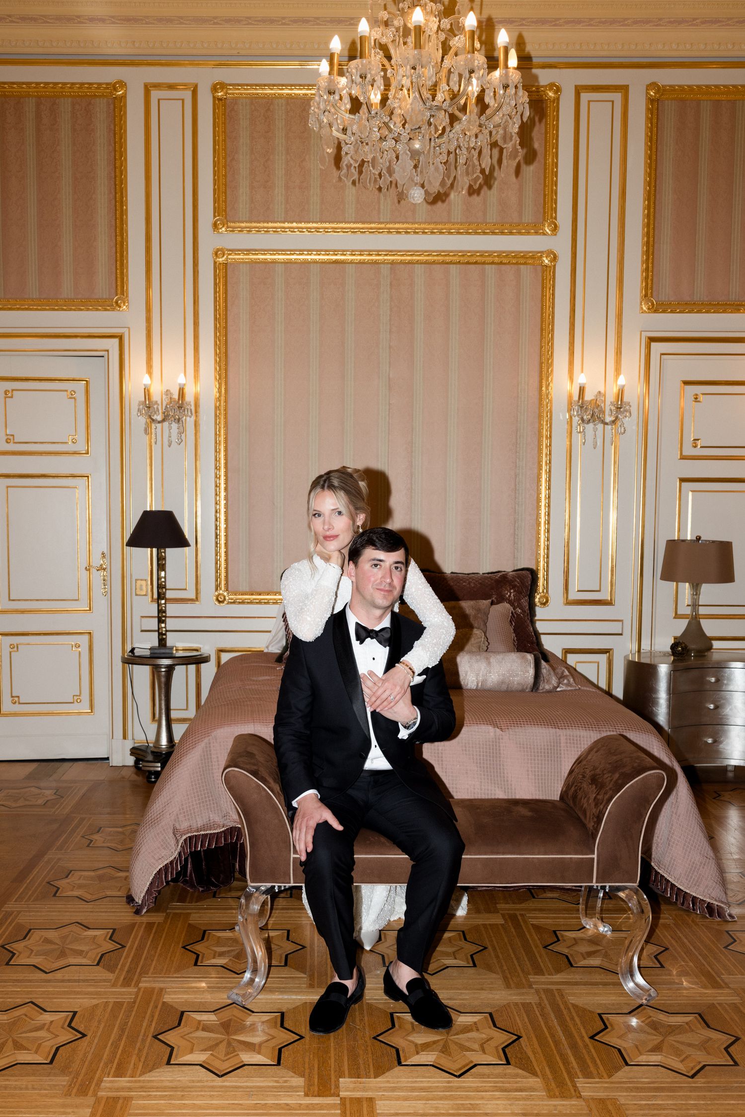 A person in formal attire sits and poses playfully on a hardwood floor in an elegant room with crystal chandeliers.