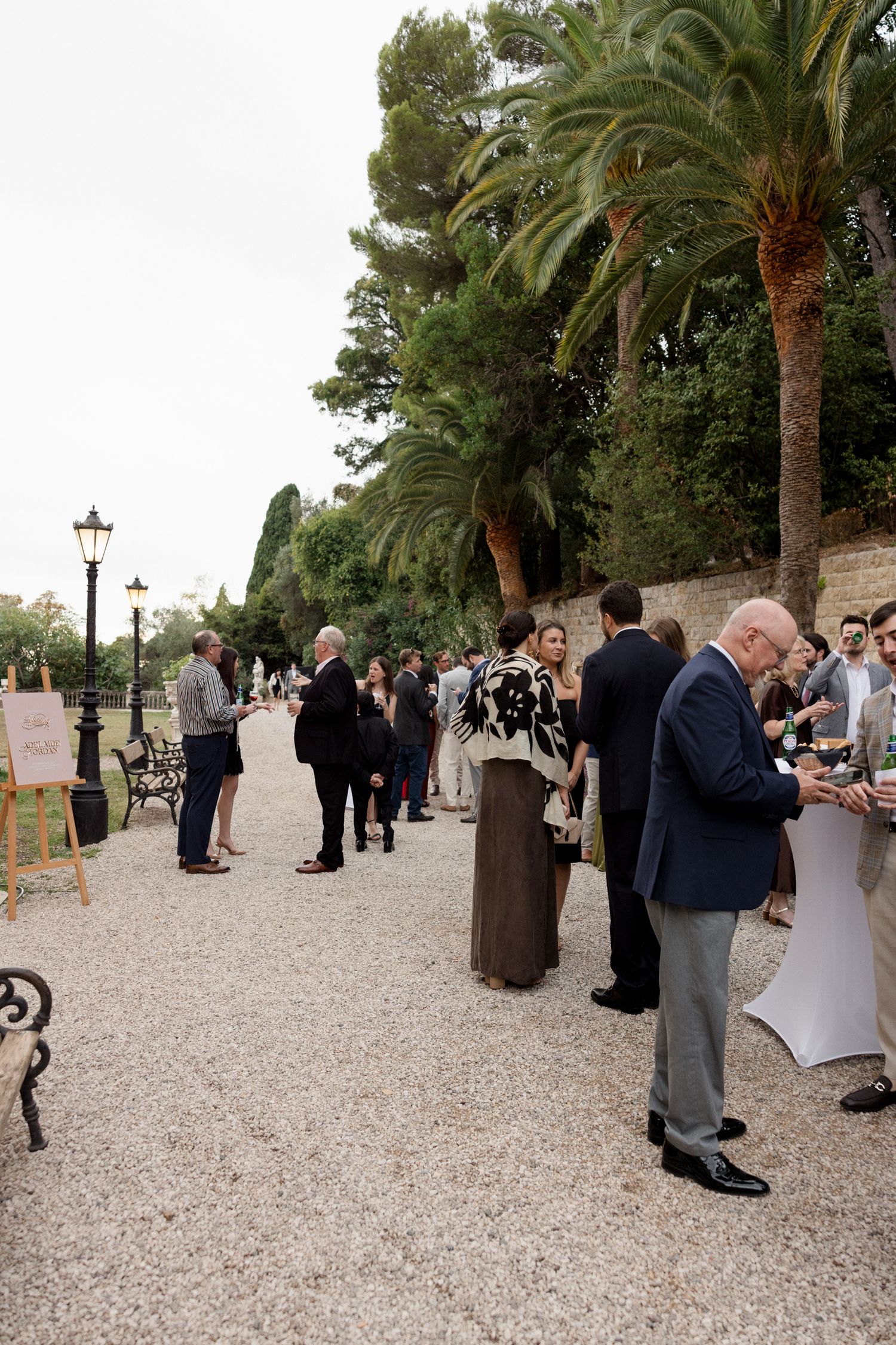 Wedding guests gather outdoors at a palm tree-lined venue with gravel paths during a cocktail reception.