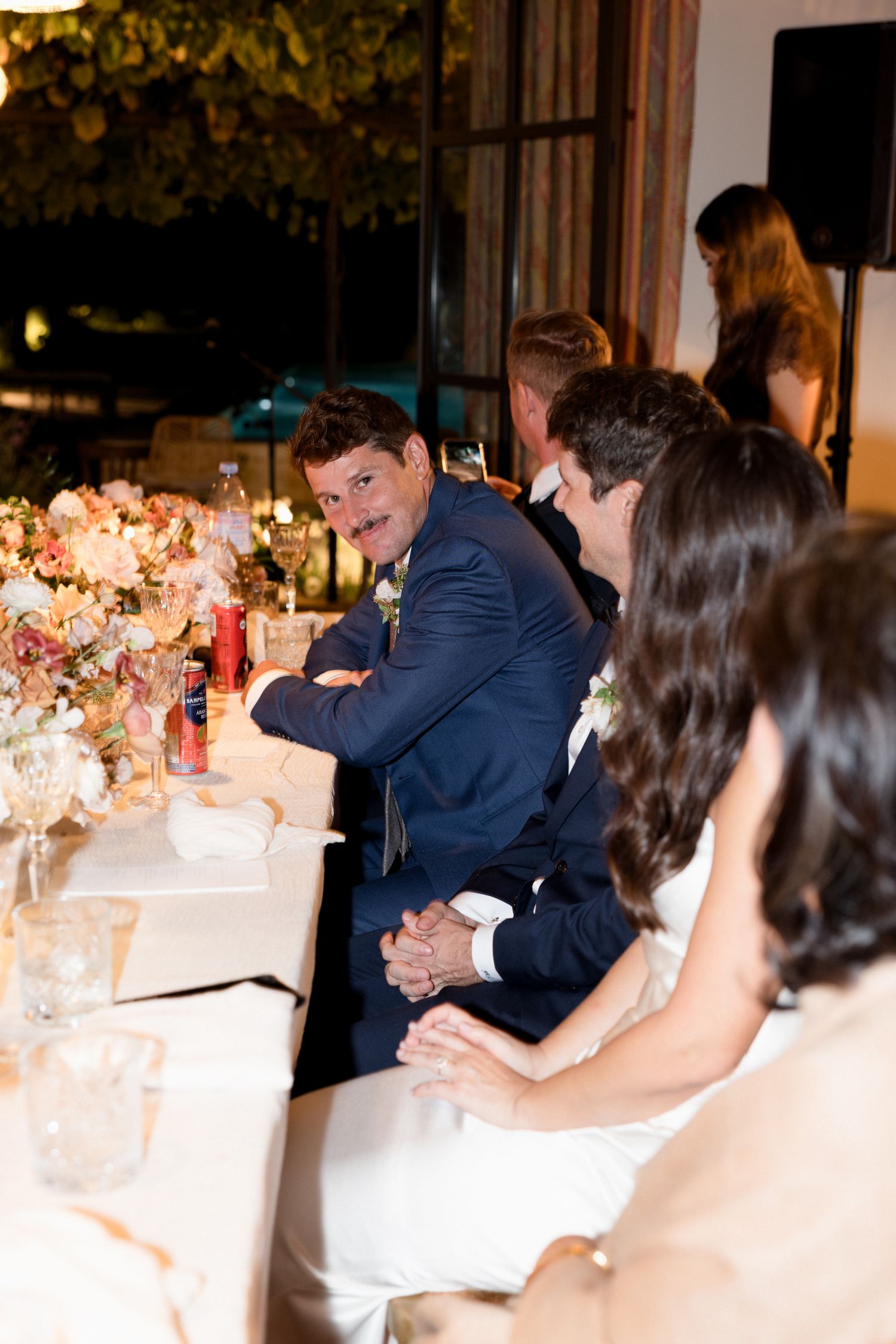 Wedding guests seated at an elegant reception table with floral centerpieces and candlelight in the evening.