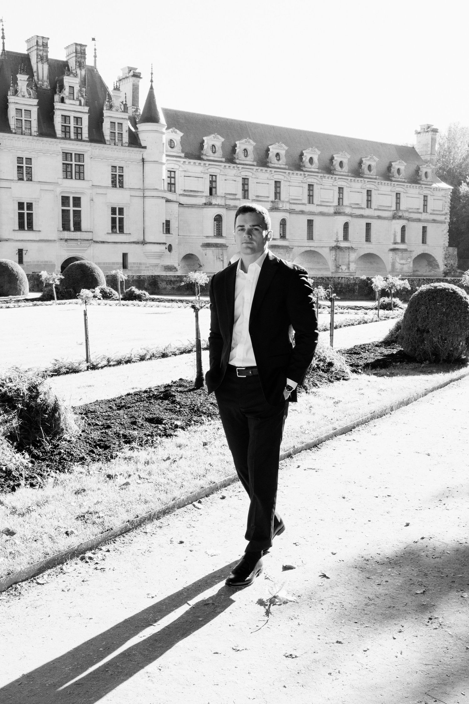 Groom in black suit walking along a sunlit garden path with Château de Chenonceau in the background.