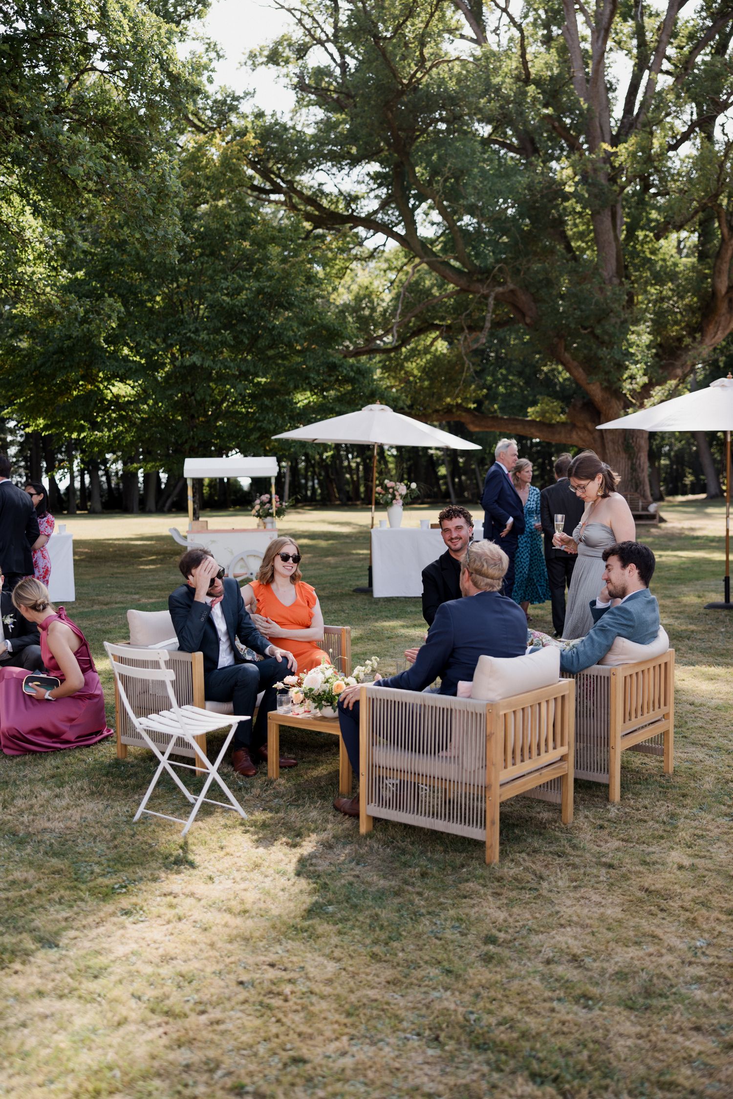 A group of people relaxing on wooden benches and chairs at an outdoor gathering in a shaded park area.