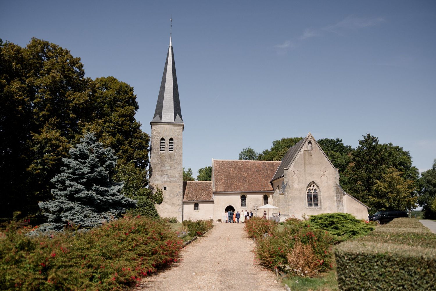 Historic stone church with tall spire nestled among trees and greenery in rural countryside setting.
