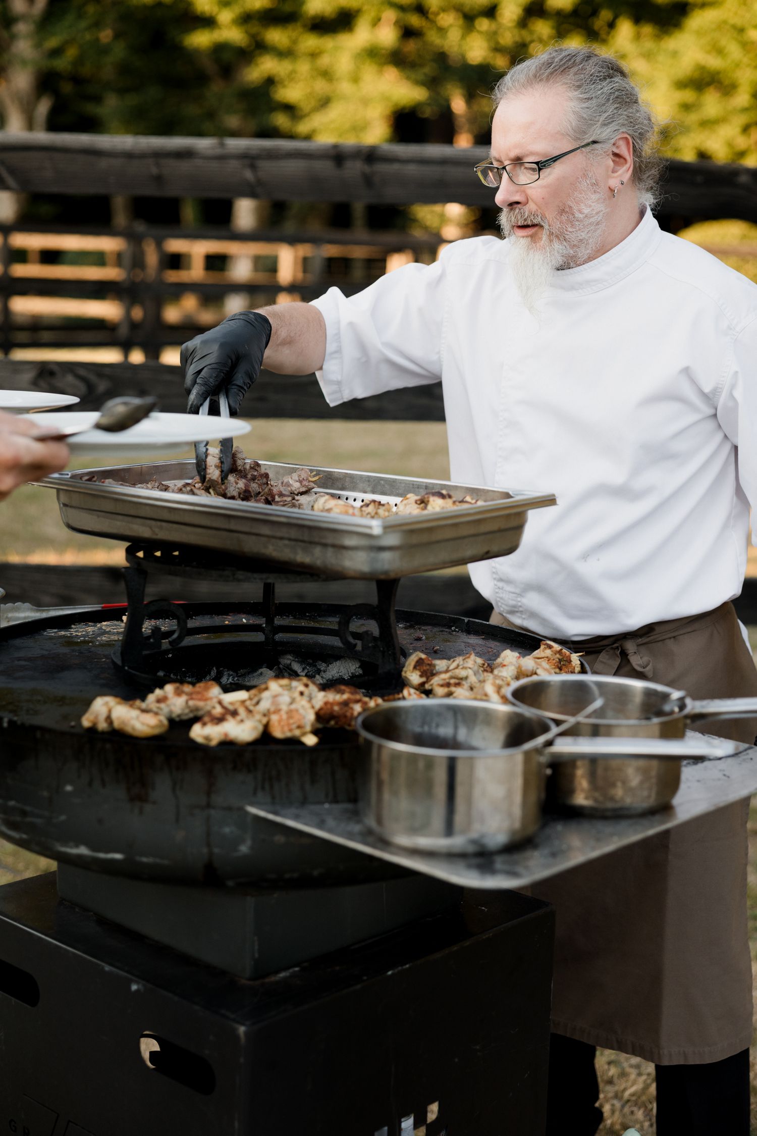 Chef in white coat grills meat on outdoor barbecue station during catering event.