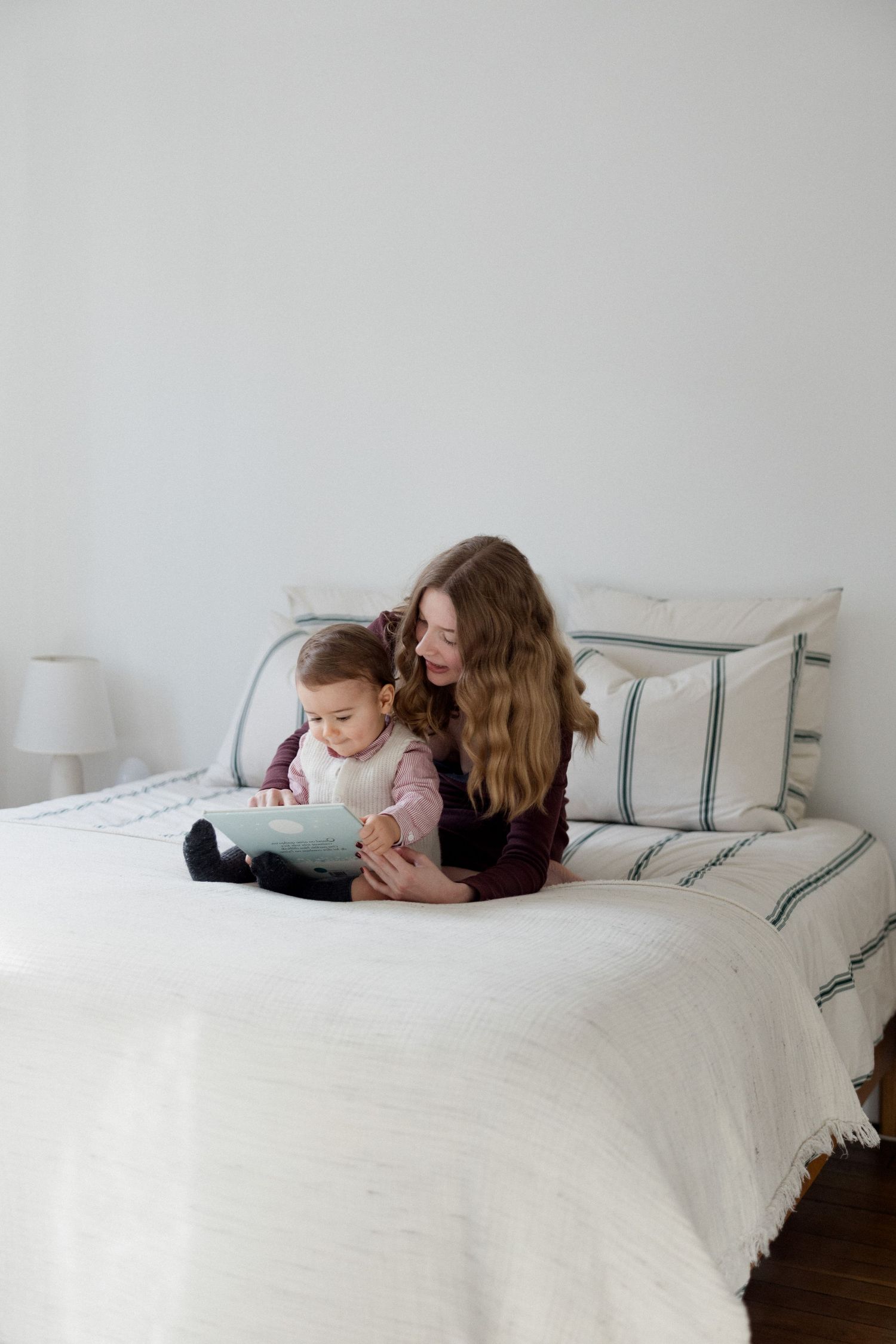 Mother with long wavy hair sitting on white bed reading to toddler in bright minimalist bedroom.