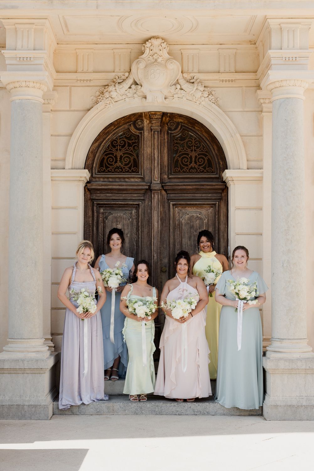 Bridesmaids wearing pastel dresses pose together in a classic stone archway at an elegant wedding venue. Château des 3 Fontaines photographe
