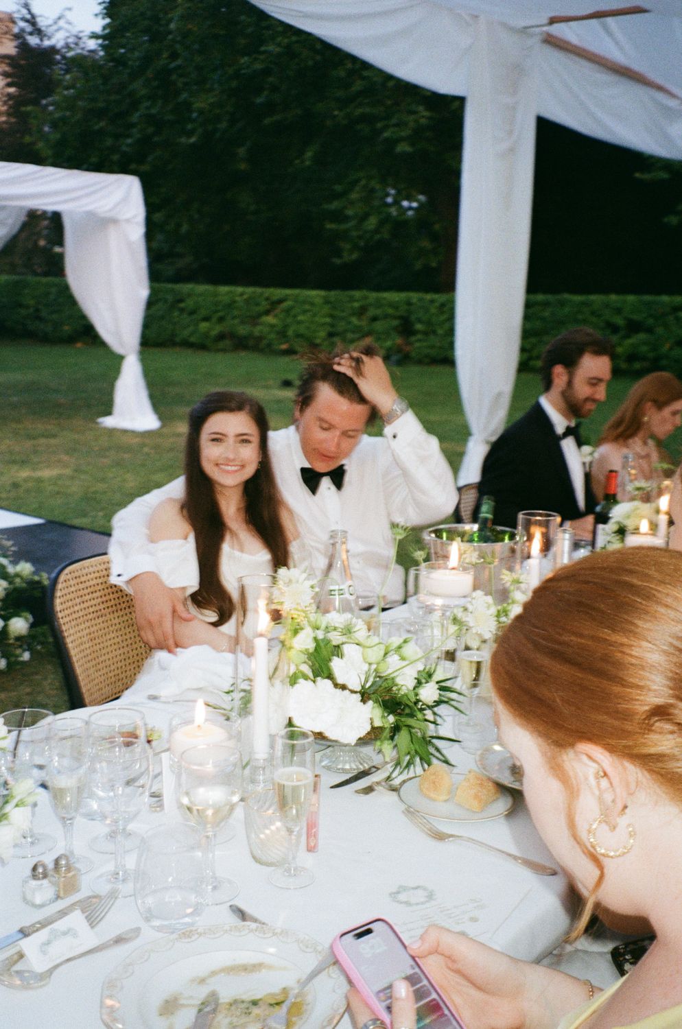 Several guests enjoy an elegant outdoor dinner reception under white drapes and floral table decor. Wedding on film