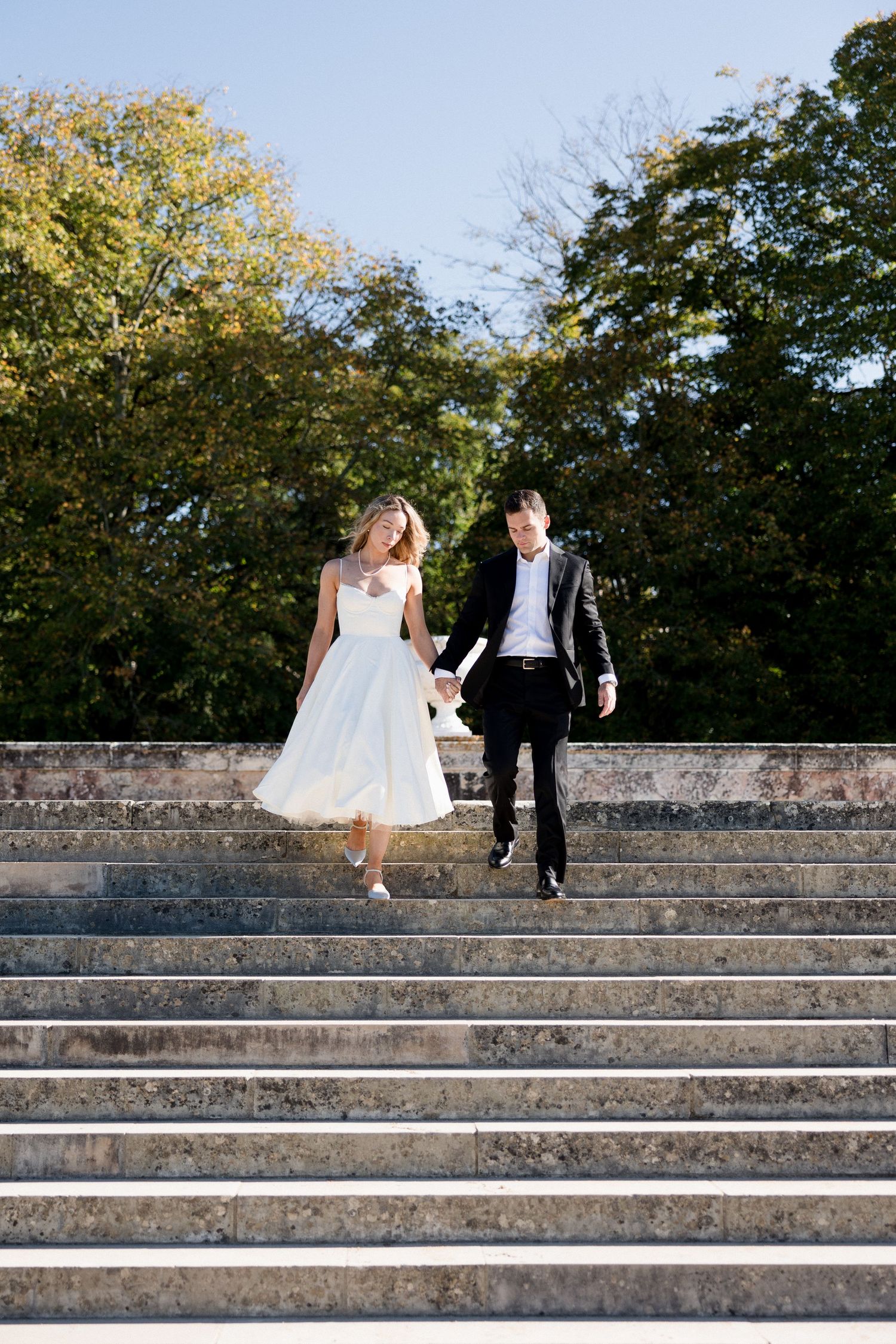 Bride and groom walking down grand stone steps hand in hand, bride in white tea-length dress outdoors.