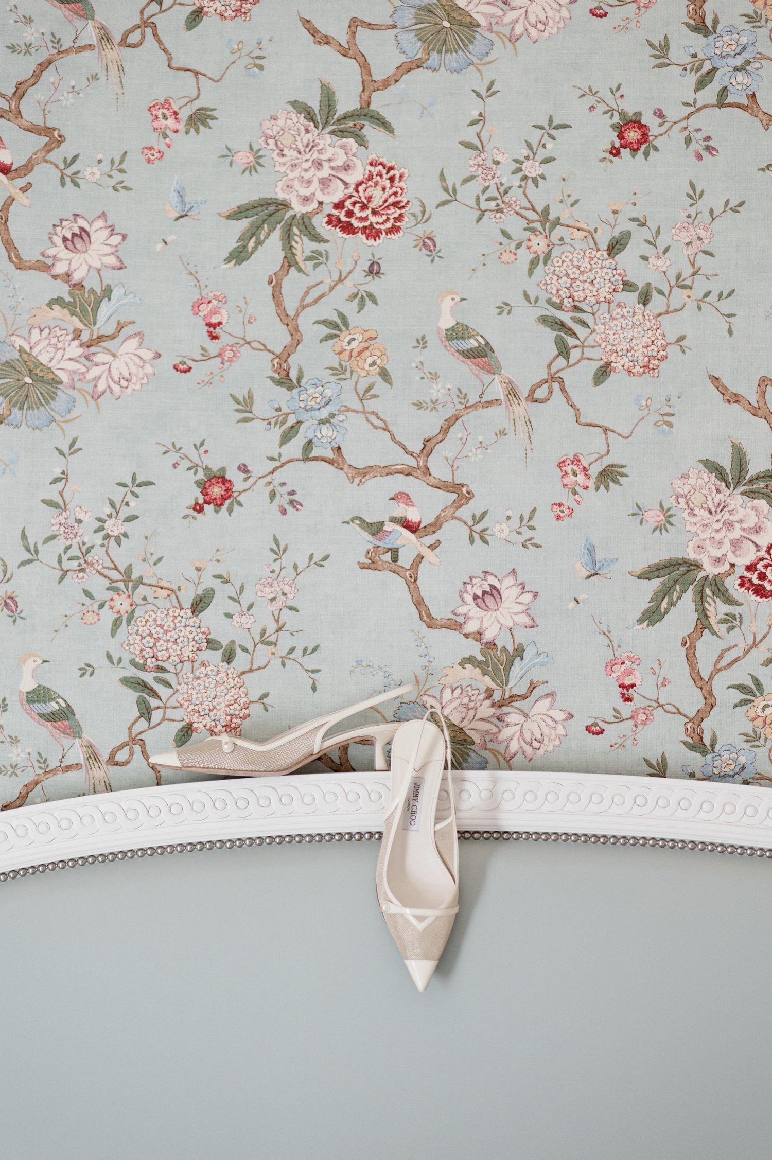 Elegant white bridal heels displayed on a mirror frame against soft blue chinoiserie floral wallpaper.