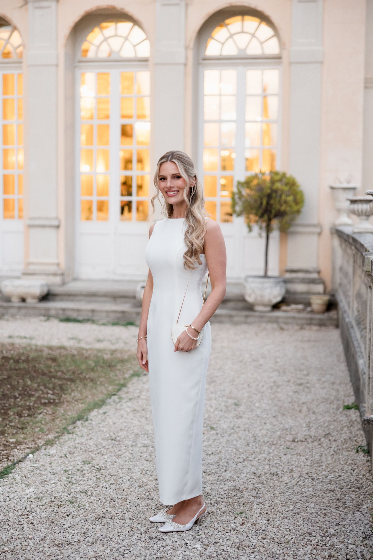Elegant white sleeveless column dress photographed against arched windows and stone architecture at sunset.
