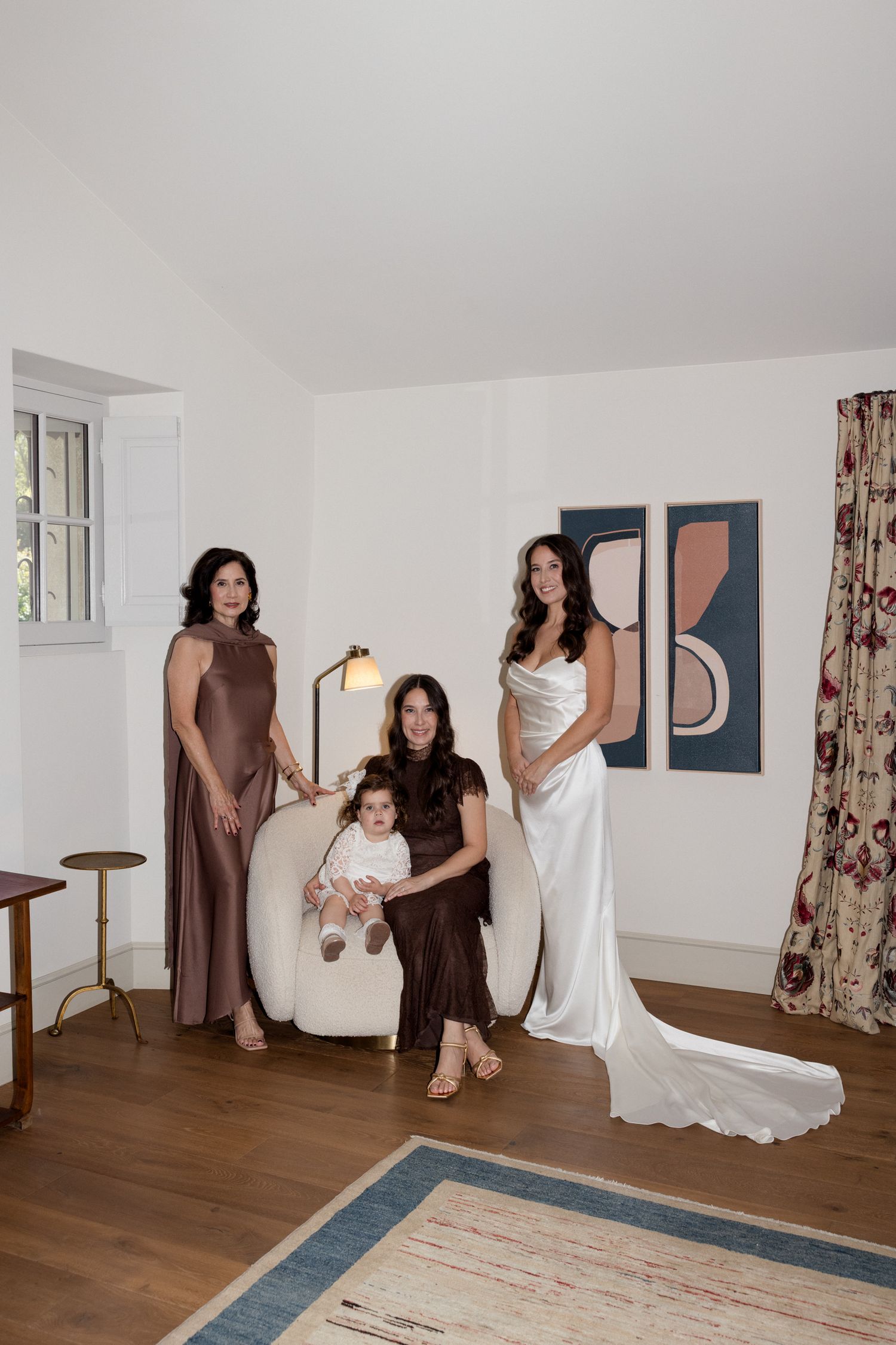 Three people in elegant dresses gather in a minimalist white room with modern artwork and hardwood floors.