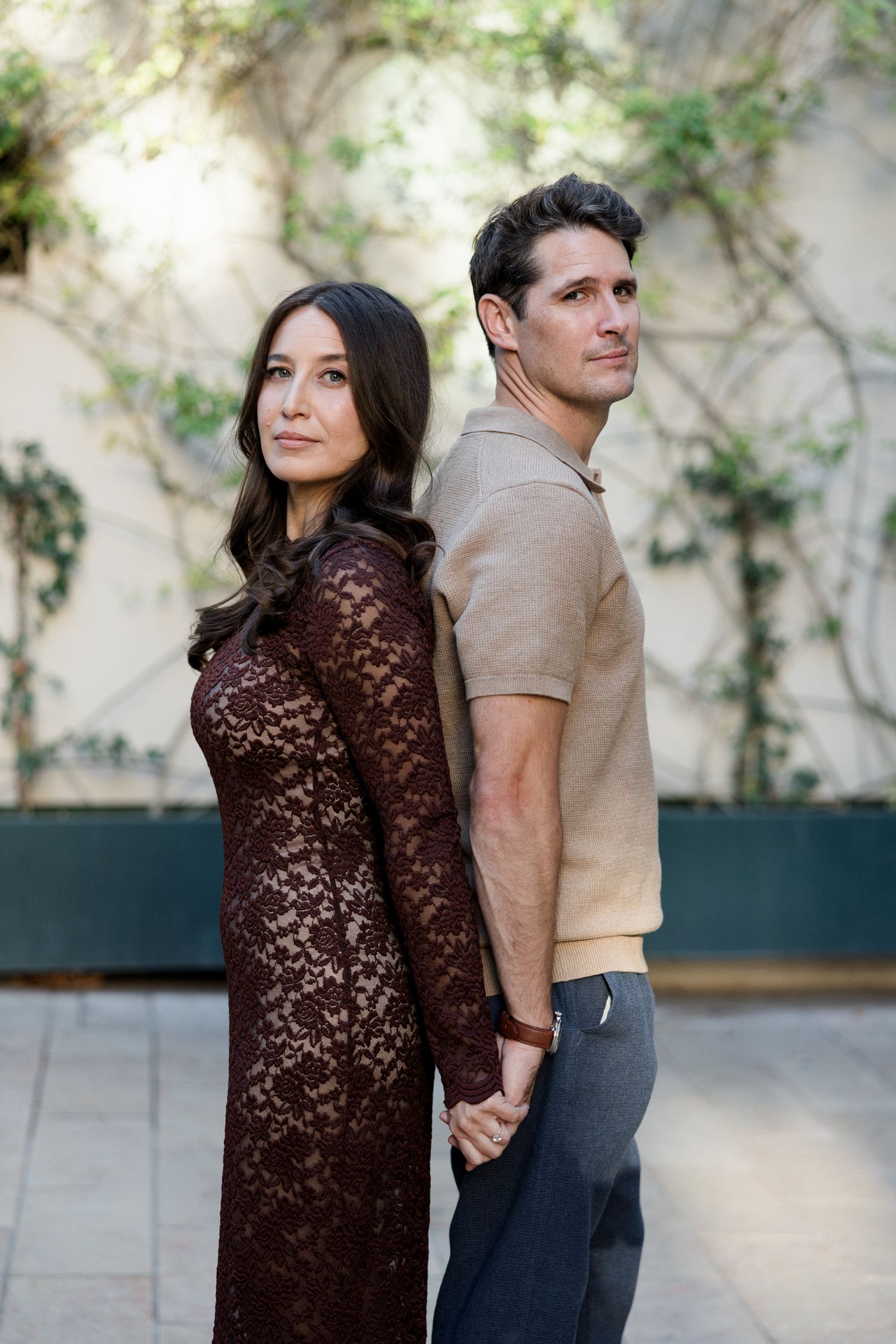 A couple in brown and beige outfits poses together against an ivy-covered wall outdoors. Engagement session France