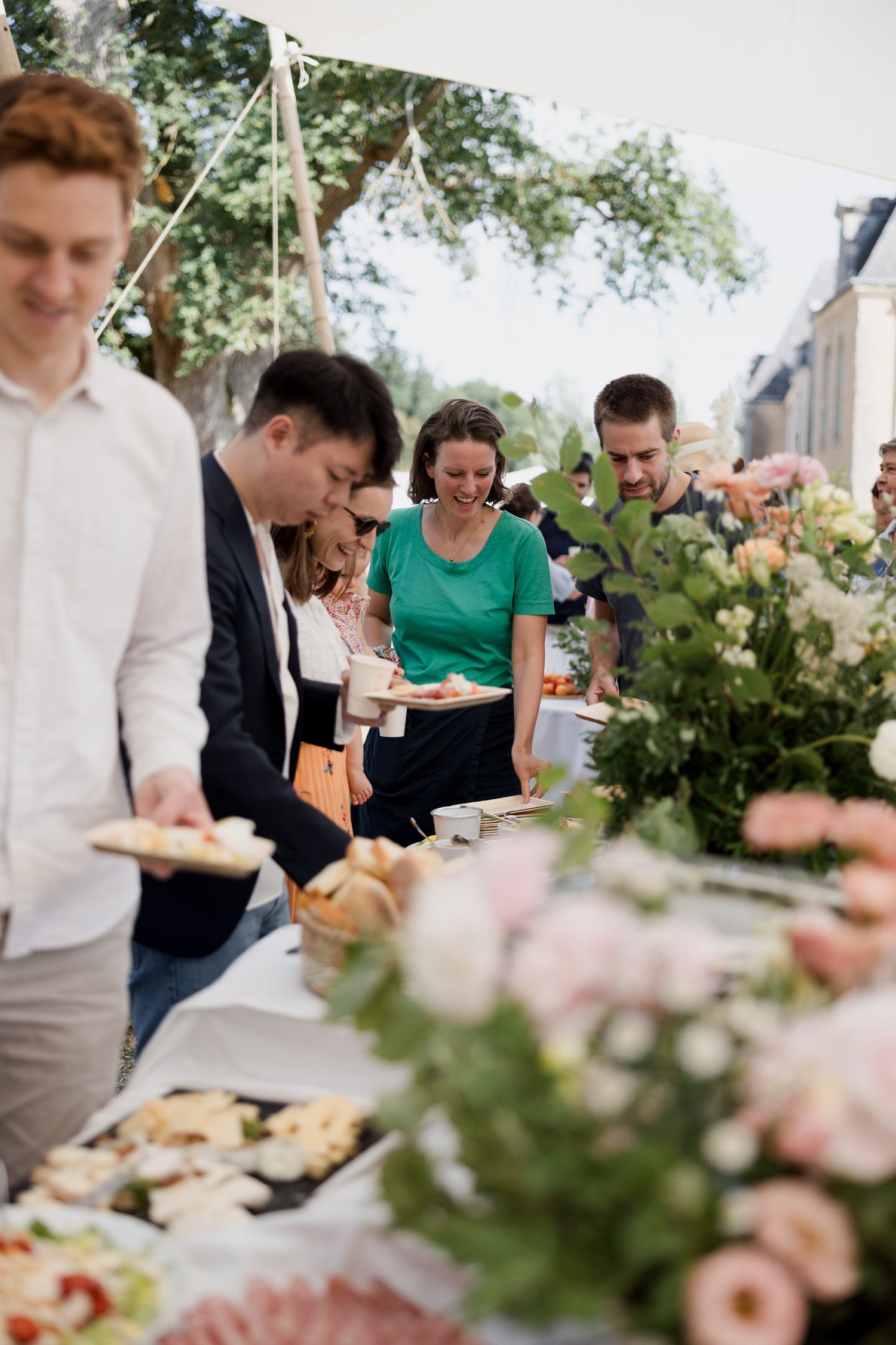 People gather around an outdoor buffet table with food and floral decorations at a casual outdoor event.
